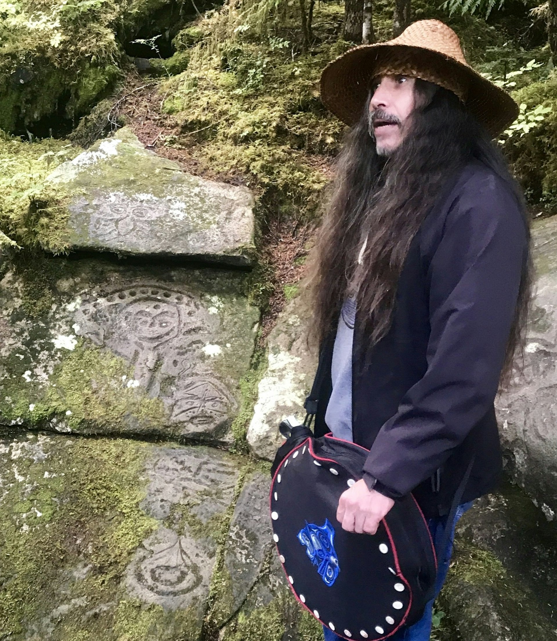 Ancient carvings etched in stone share space with green moss as a man with a drum and reed hat tells stories nearby.