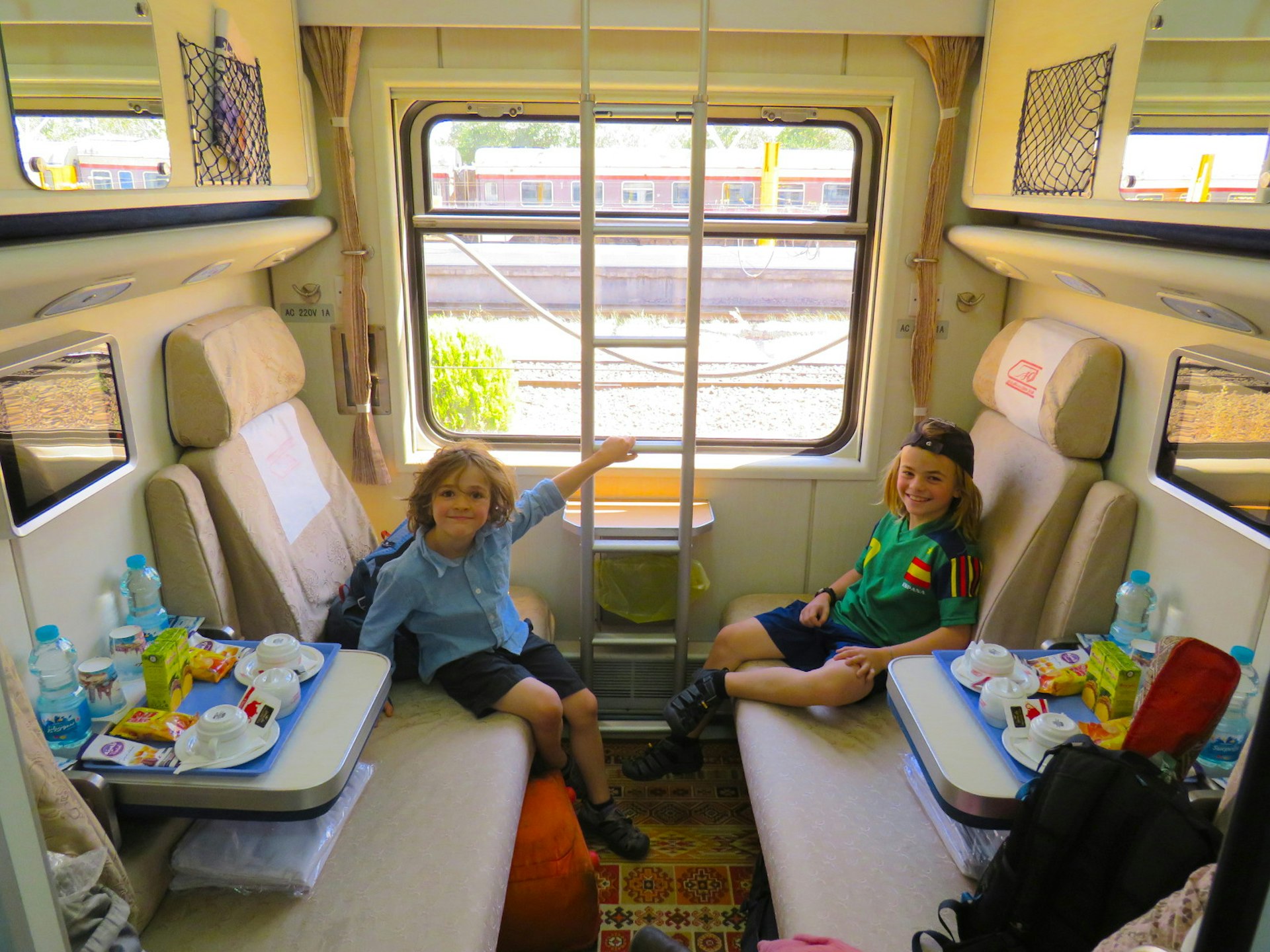 Two young boys sit in a compartment on a train, smiling at the camera