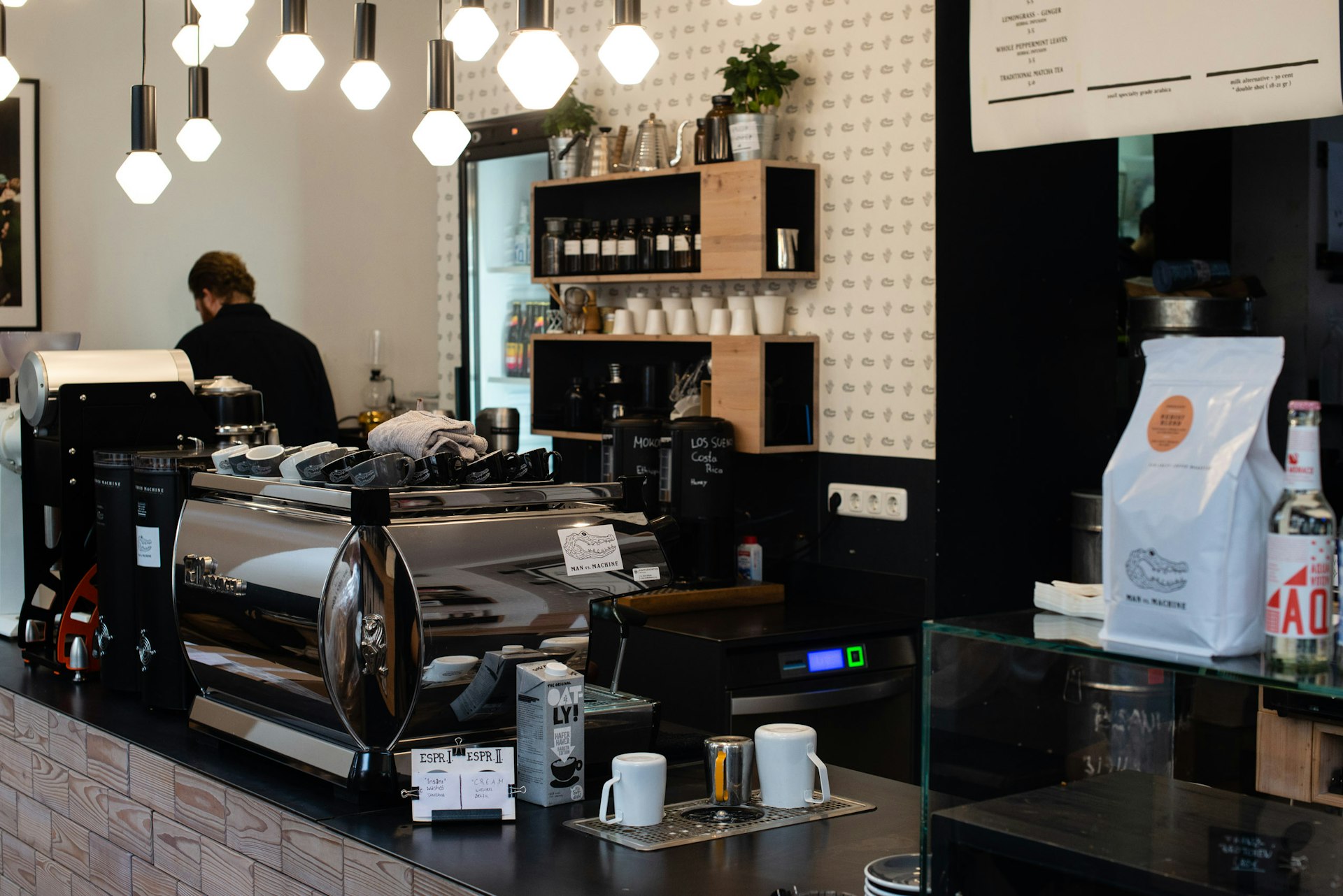 The stylish interior of Man Versus Machine cafe in Munich. A barista faces away from the camera, surrounded by gleaming chrome coffee makers and appliances. Funky, geometric light features hang from the roof. © Kate Mann / Lonely Planet