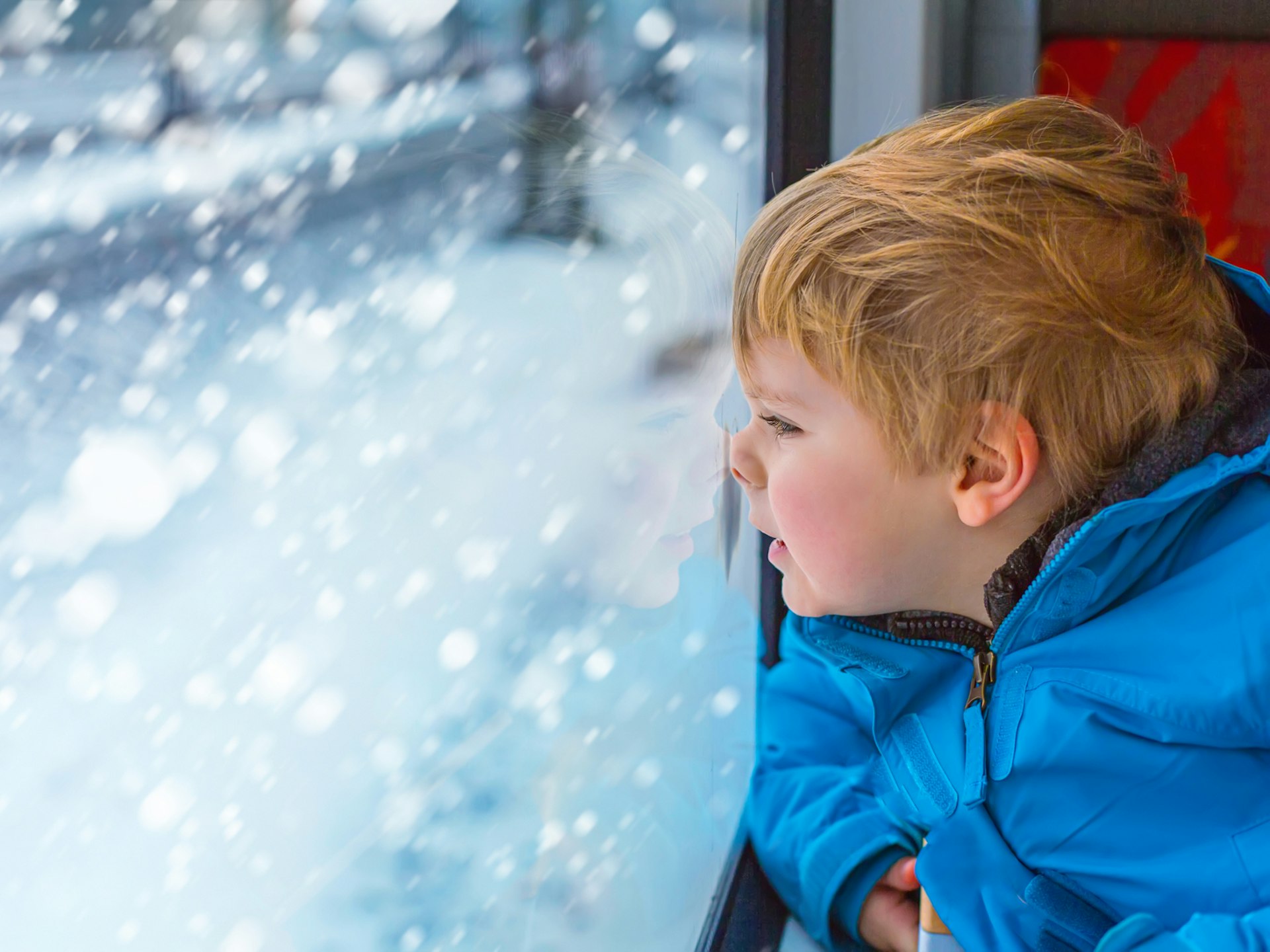 A young boy with his face pressed up against a train window watching snow fall outside