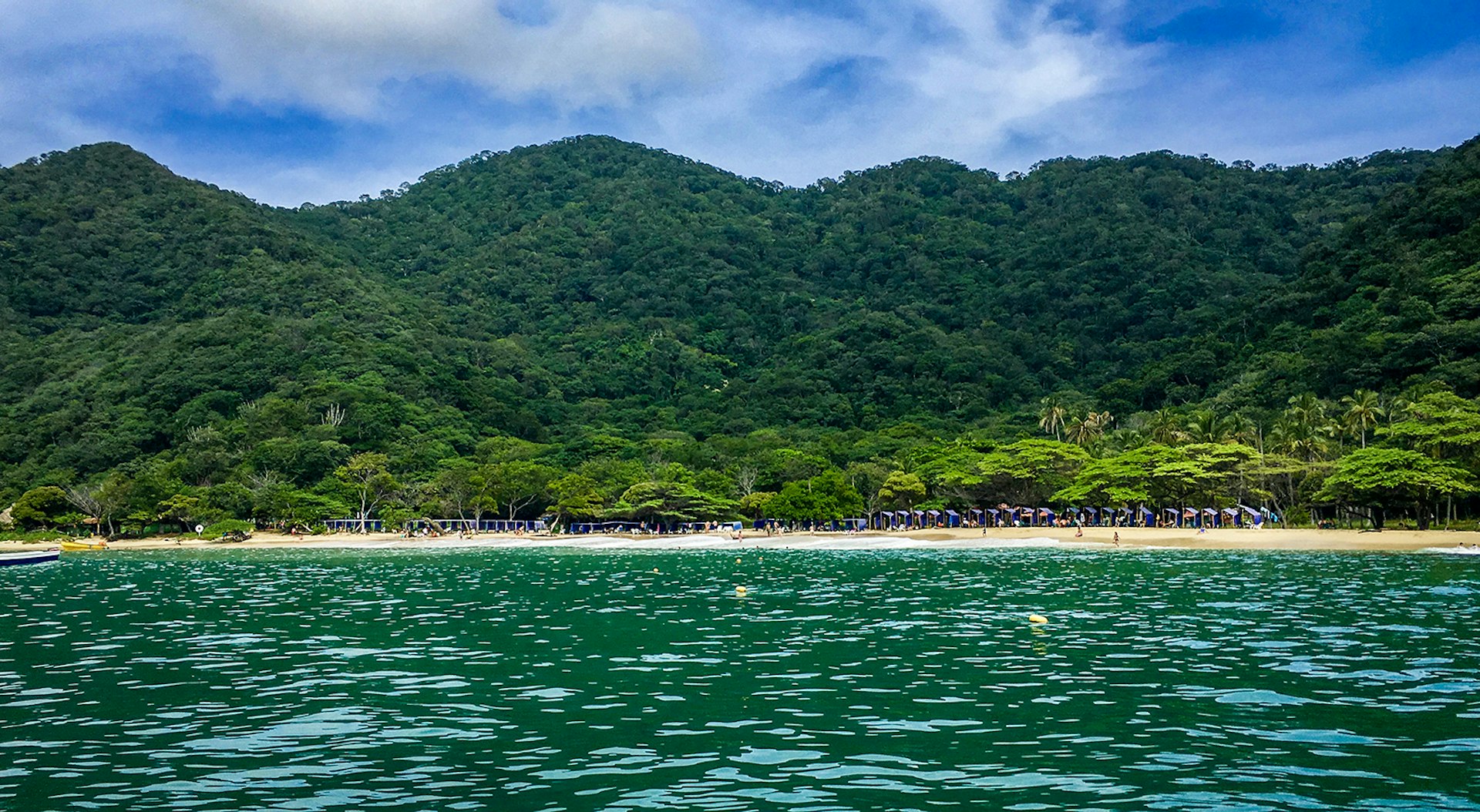 An image taken from the water of a beach backed by forested green hills