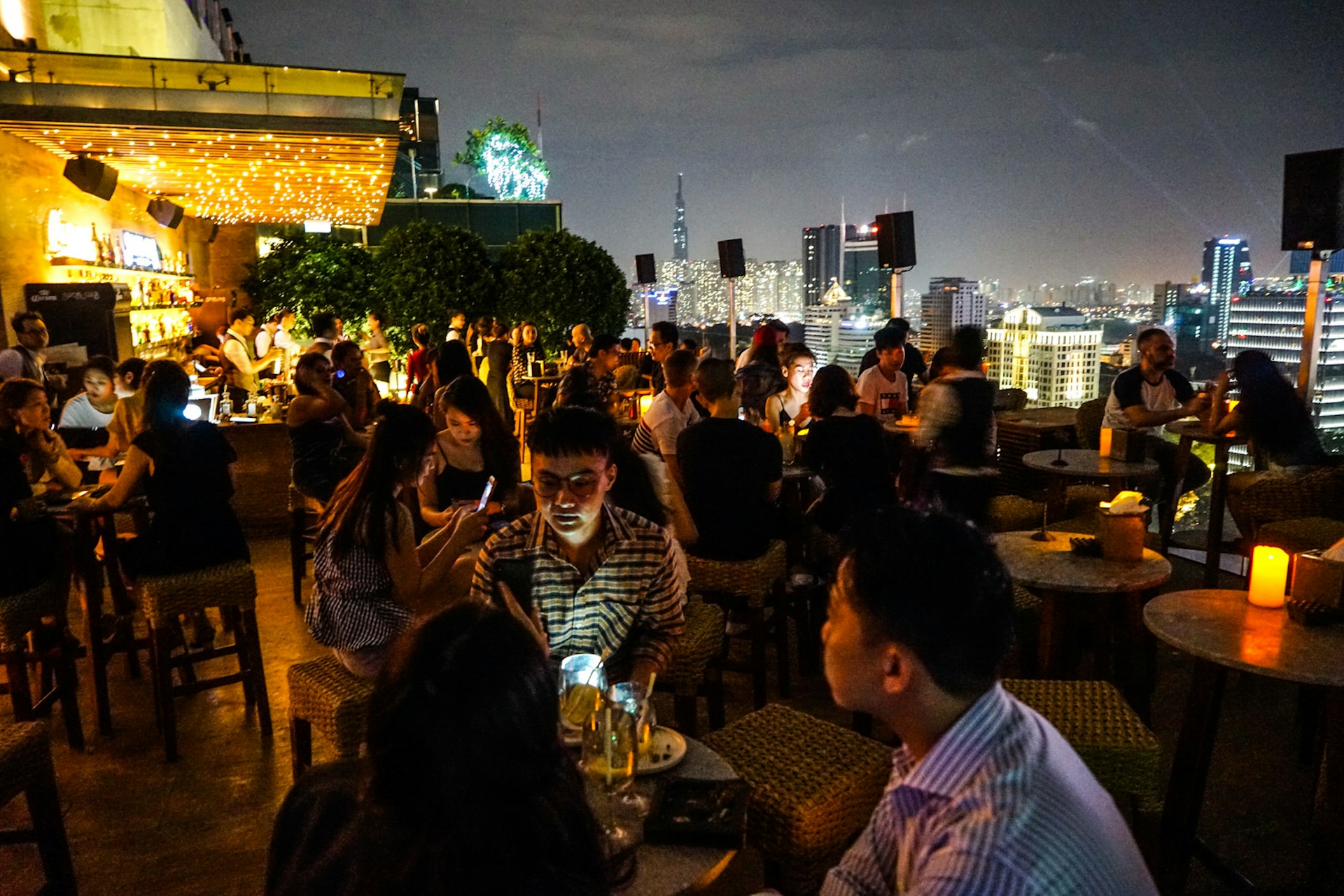 Revellers gather at Saigon Social Club's rooftop bar to drink and dine. The space twinkles with candles and fairylights as well as city lights in the background. 