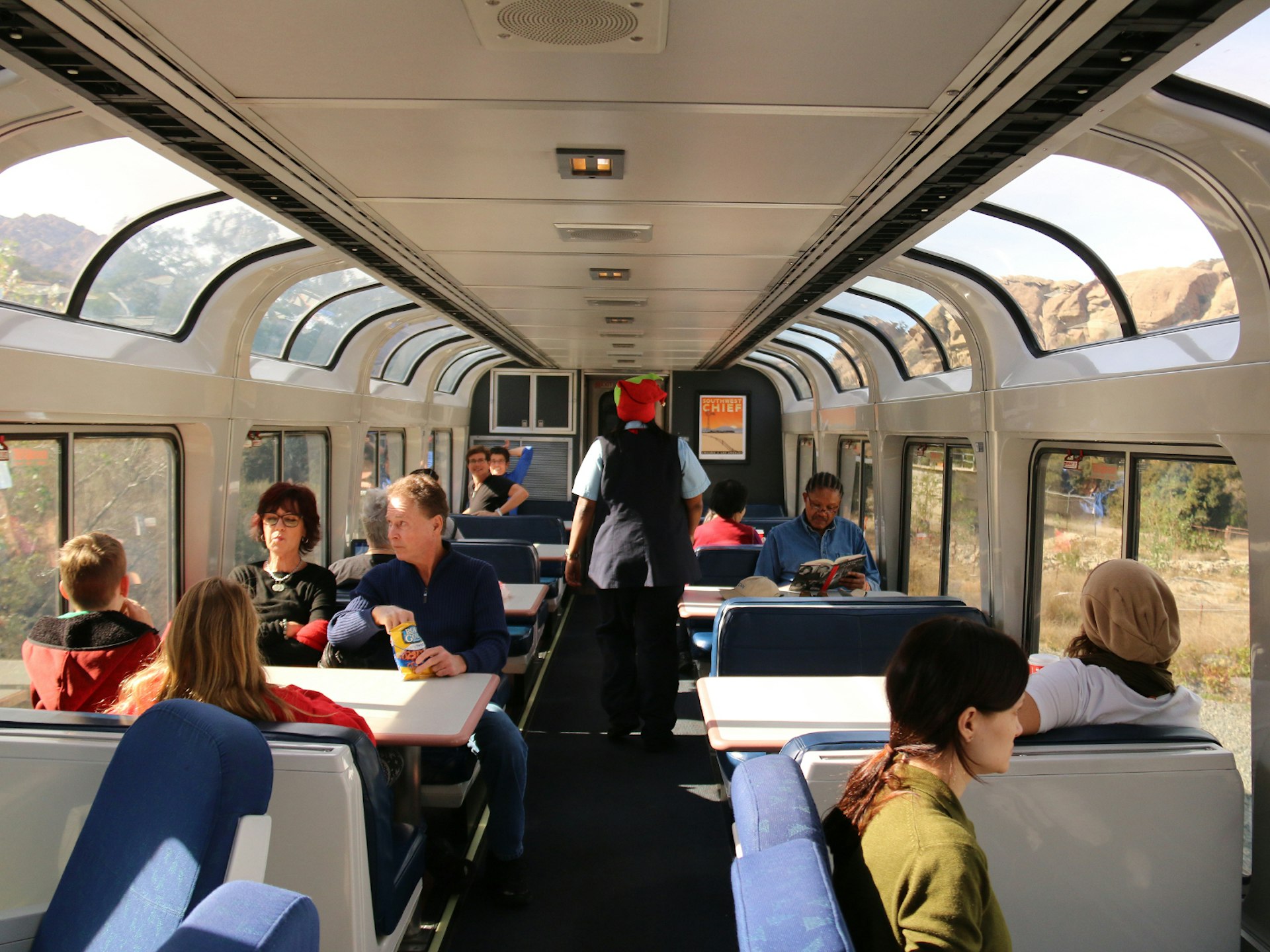 Passengers seated in a carriage of an Amtak train while an attendant stands in the middle aisle