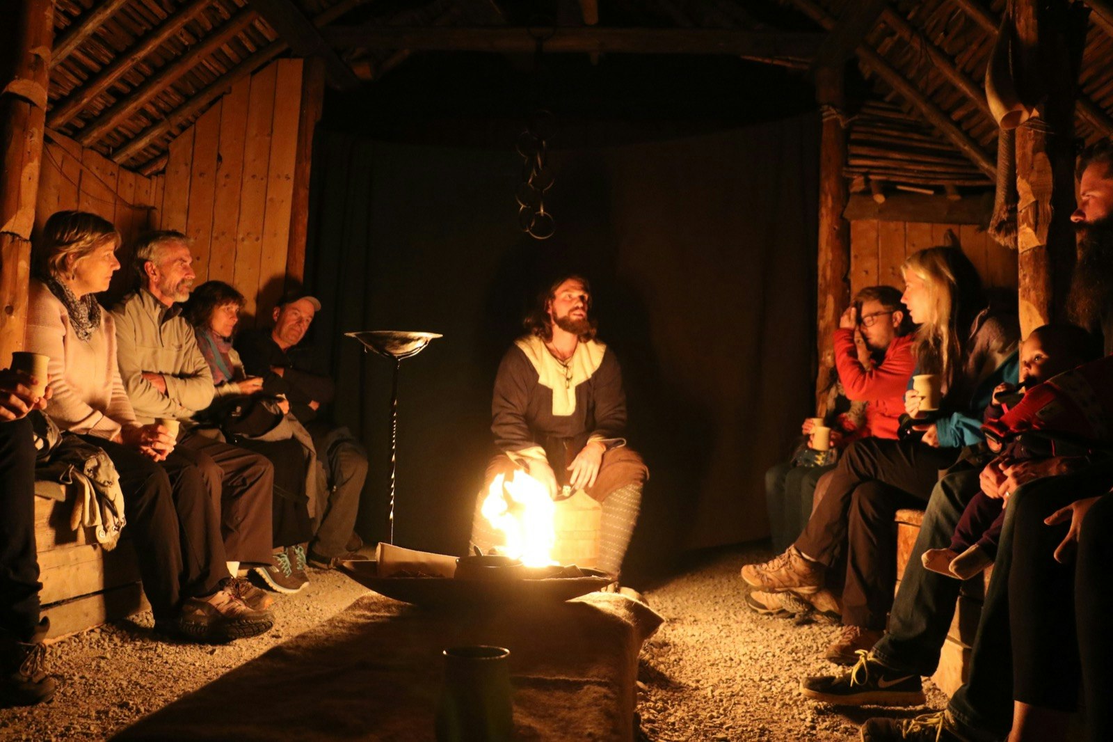 A man dressed in Viking regalia speaks to a group of travelers inside a wooden structure, illuminated by a central fire