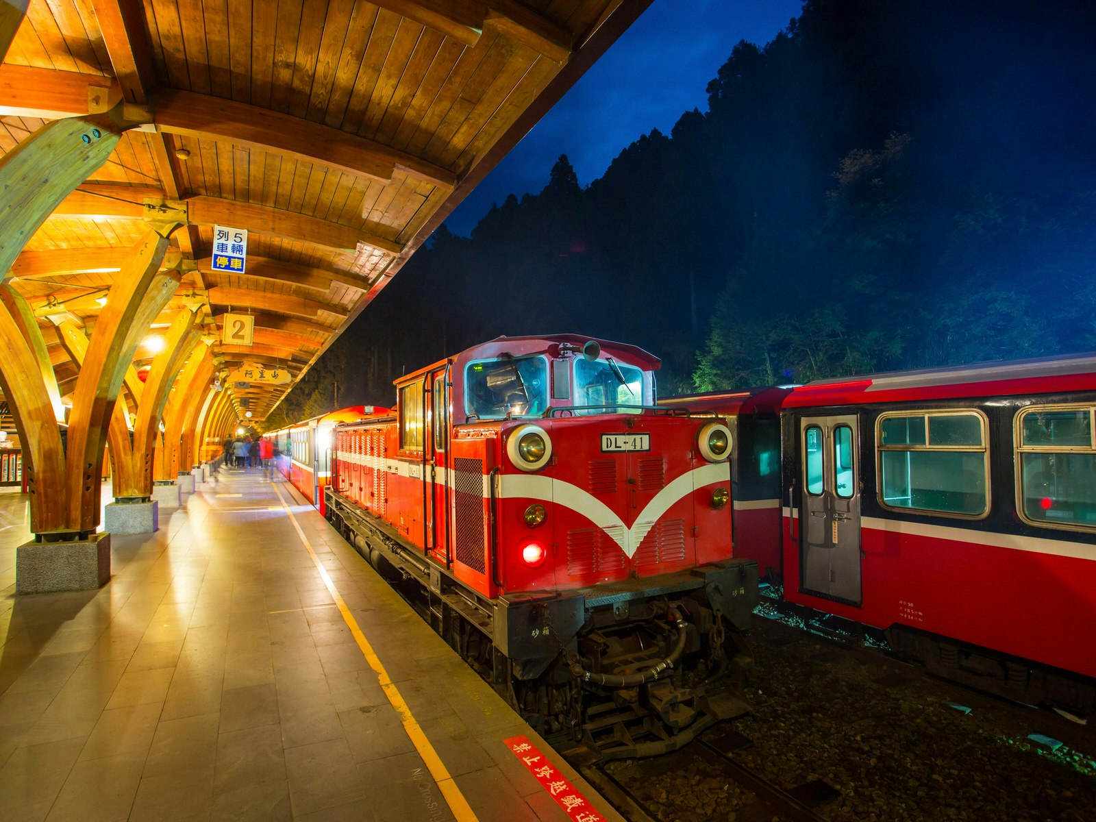 A red and white train engine parked on a train platform in the dark, with illuminated platform lights and wooden overhang.