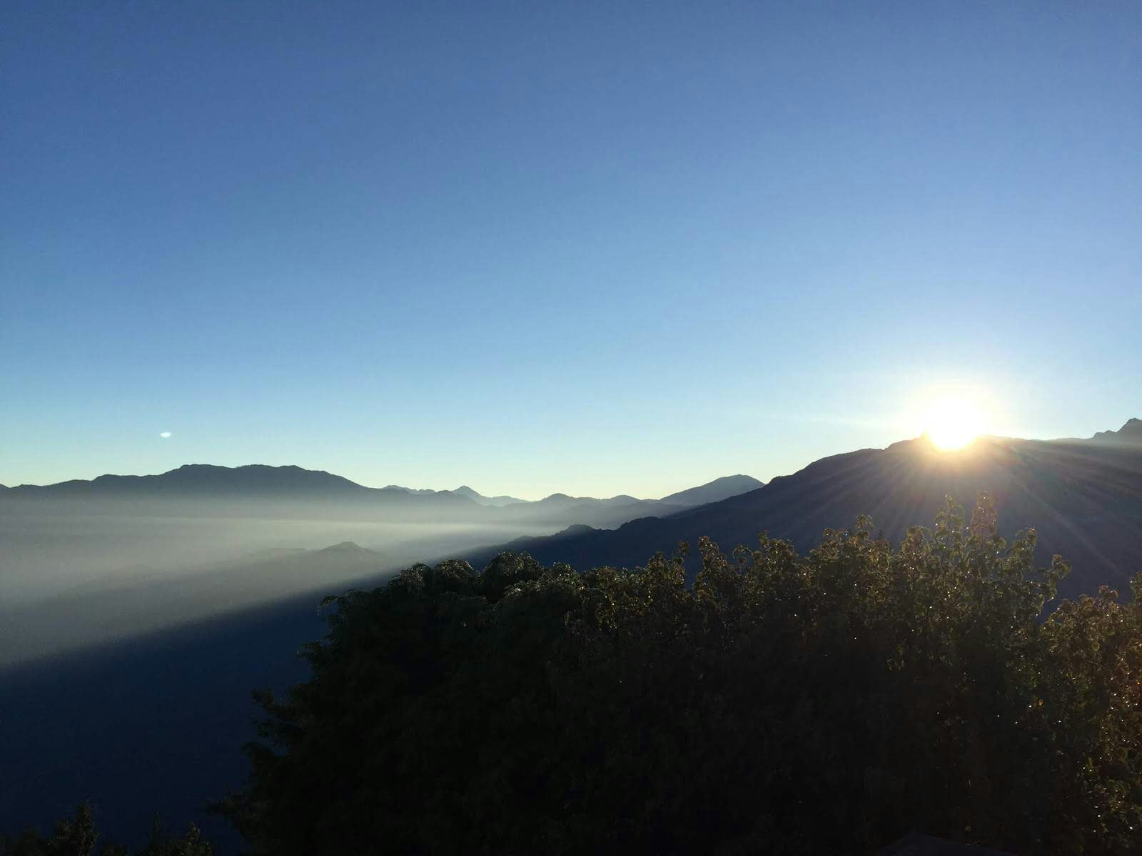 Trees in the foreground with mountains and clouds and a bright sunburst in the background.