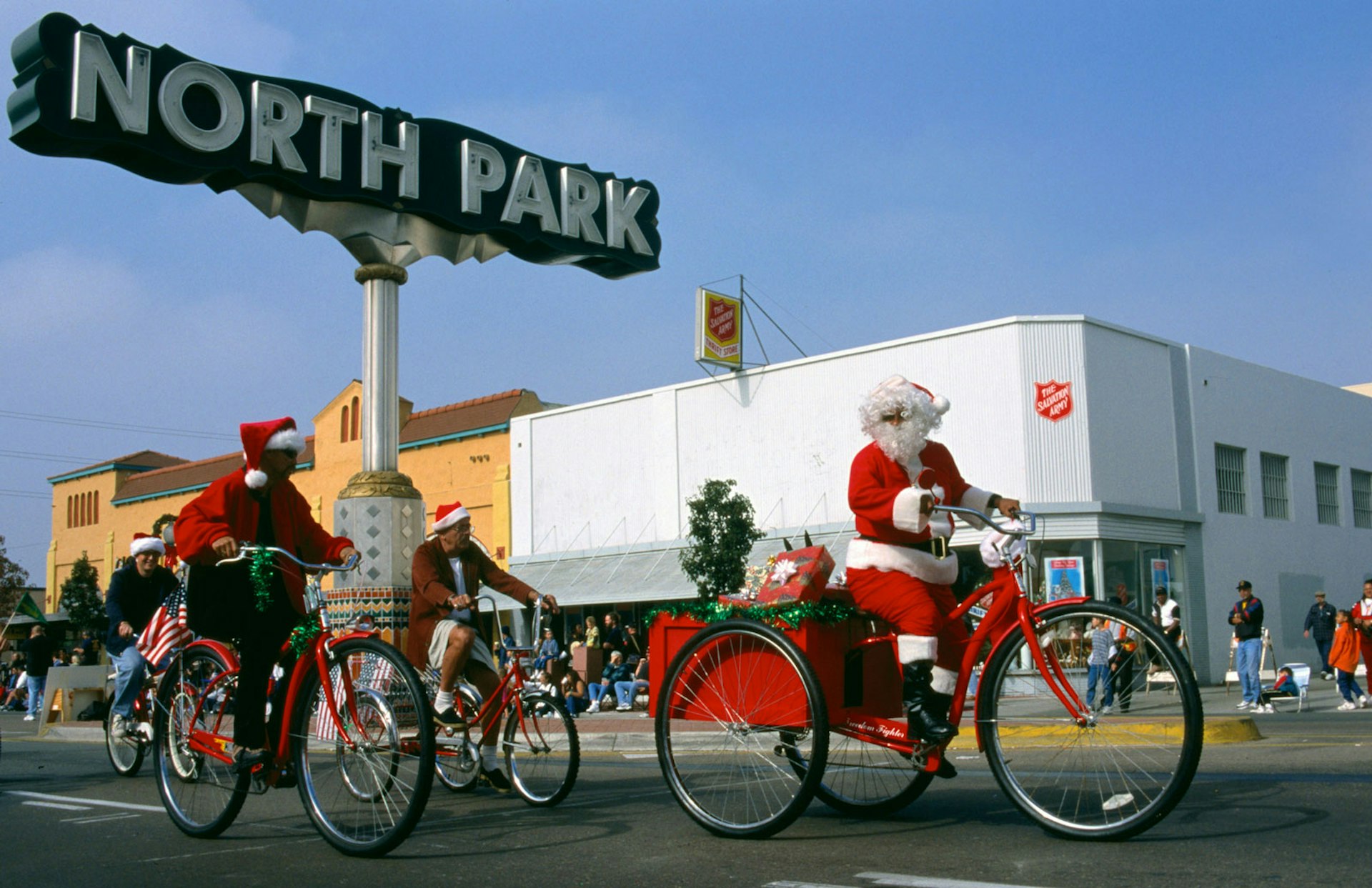 Mad dressed as Santa Clause rides a beach cruiser with a trailer filled with presents along side other men in santa hats also riding bikes