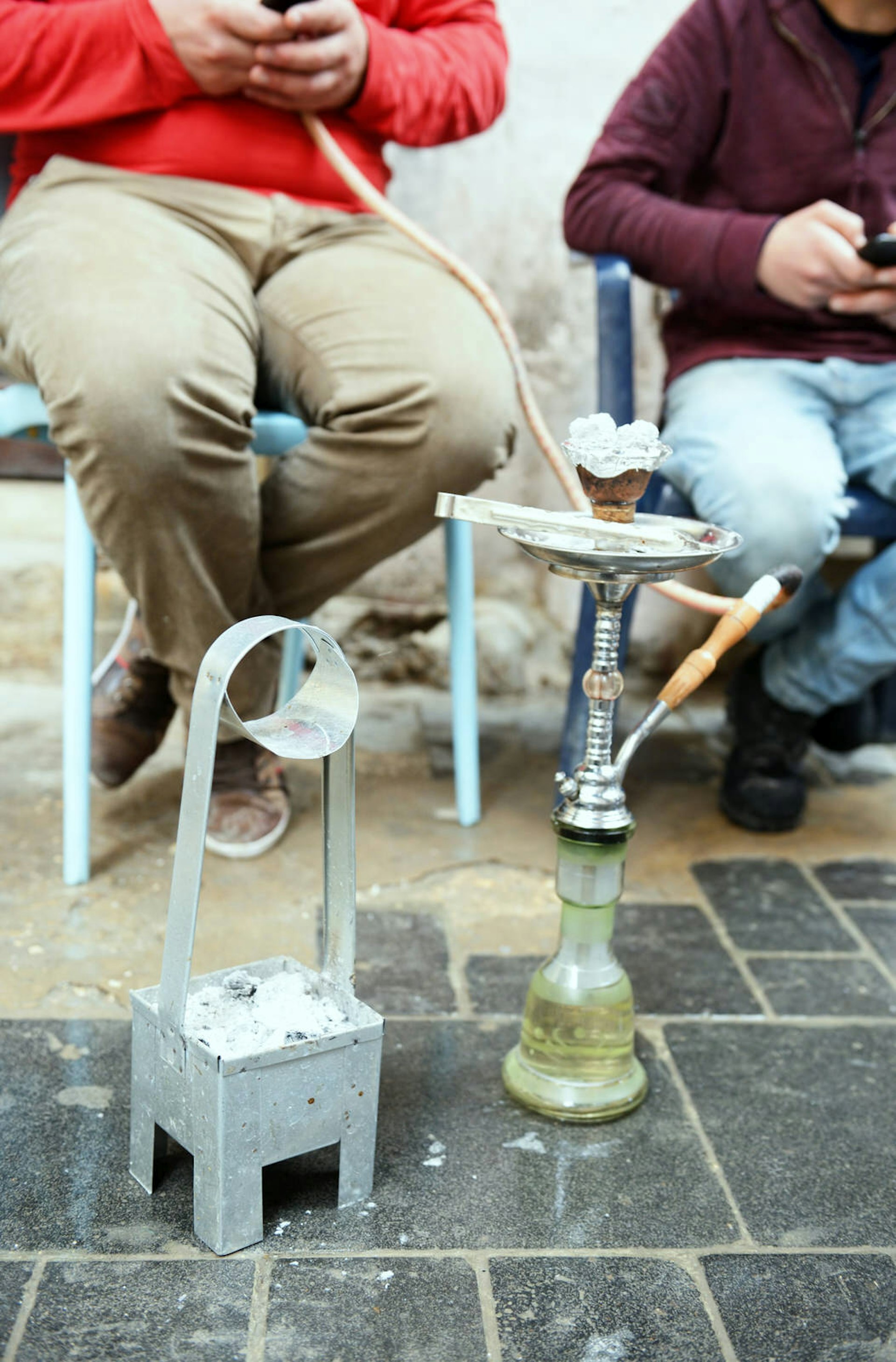 A hookah, a traditional Middle Eastern, device for smoking tobacco, sits on a sidewalk in Lebanon, with two smokers in chairs behind
