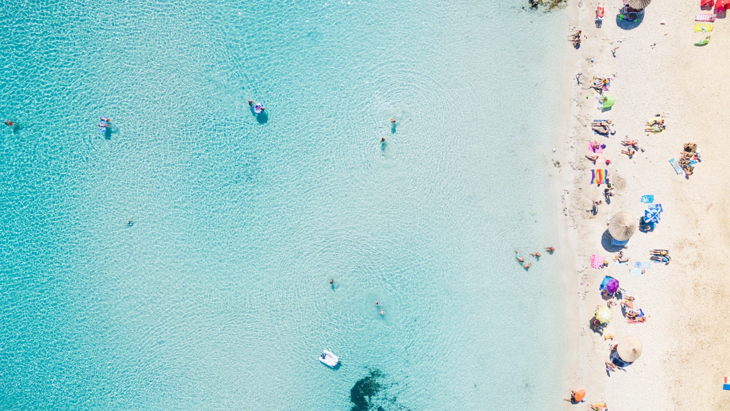 Aerial of a beach with tourists swimming in clear water and lounging on the sand.