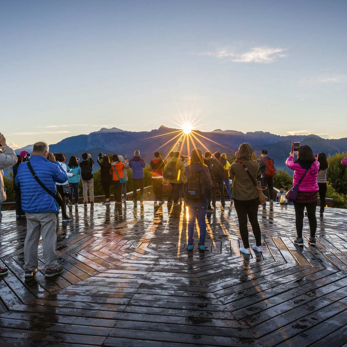 A group of tourists point cameras toward the sun rising over a line of mountain peaks.