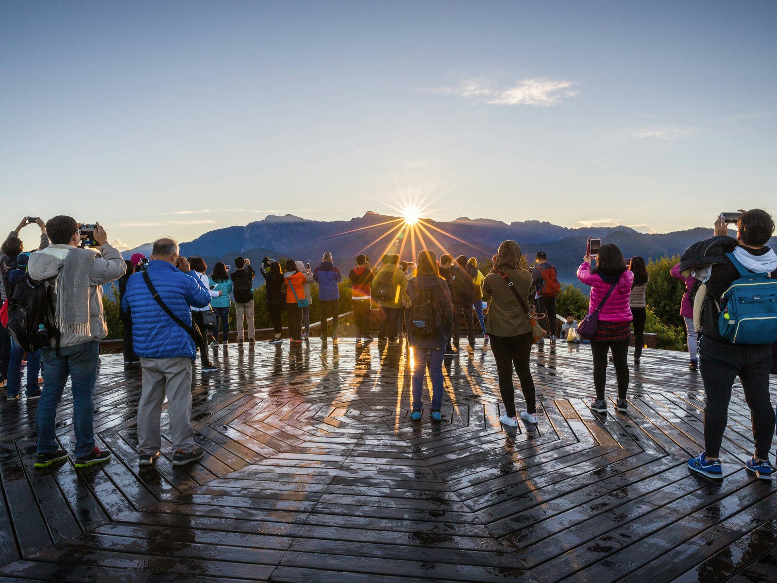 A group of tourists point cameras toward the sun rising over a line of mountain peaks.