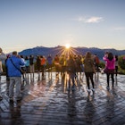 A group of tourists point cameras toward the sun rising over a line of mountain peaks.