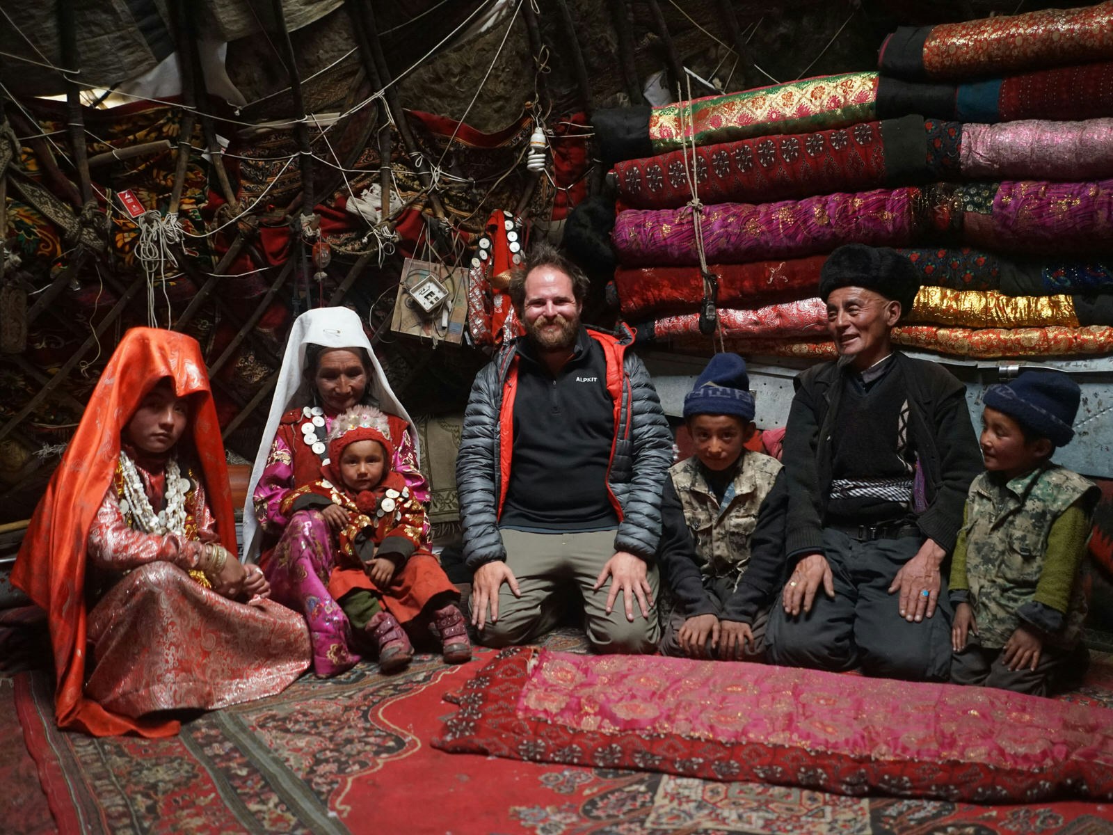 Writer Jonny Duncan kneels with a Kyrgyz nomad family inside their yurt, which is draped with red textiles and carpets