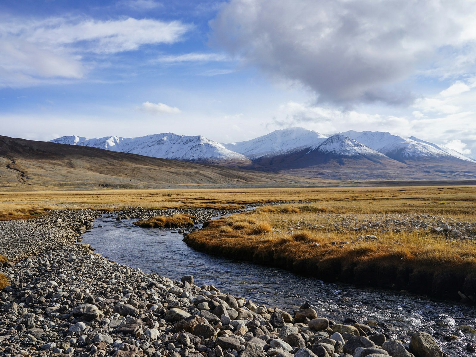 View of a river in the foreground with yellowed grass meadow and snow-capped mountains in background