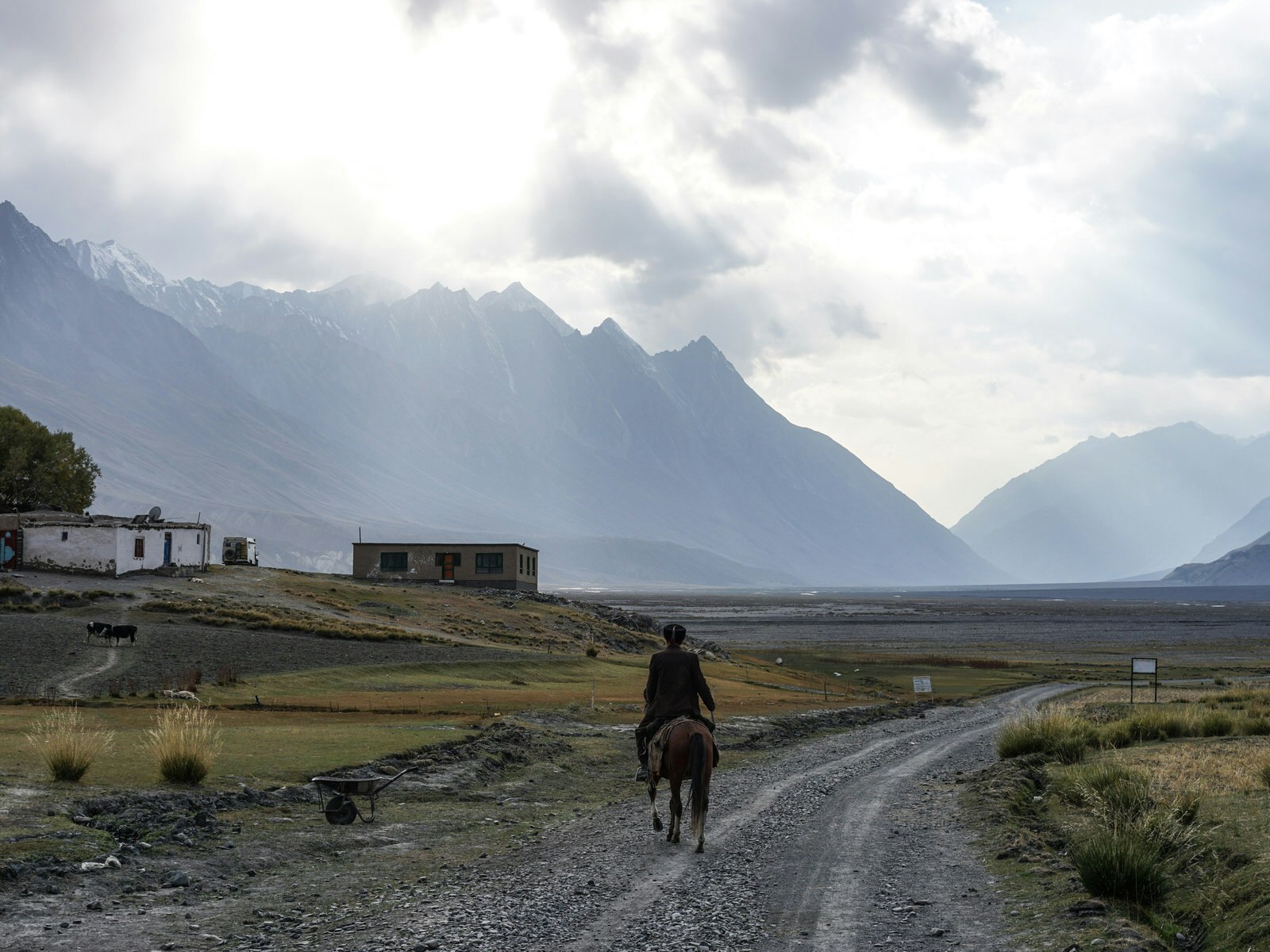 A man rides a donkey down a desolate dirt road with huge mountains in the background