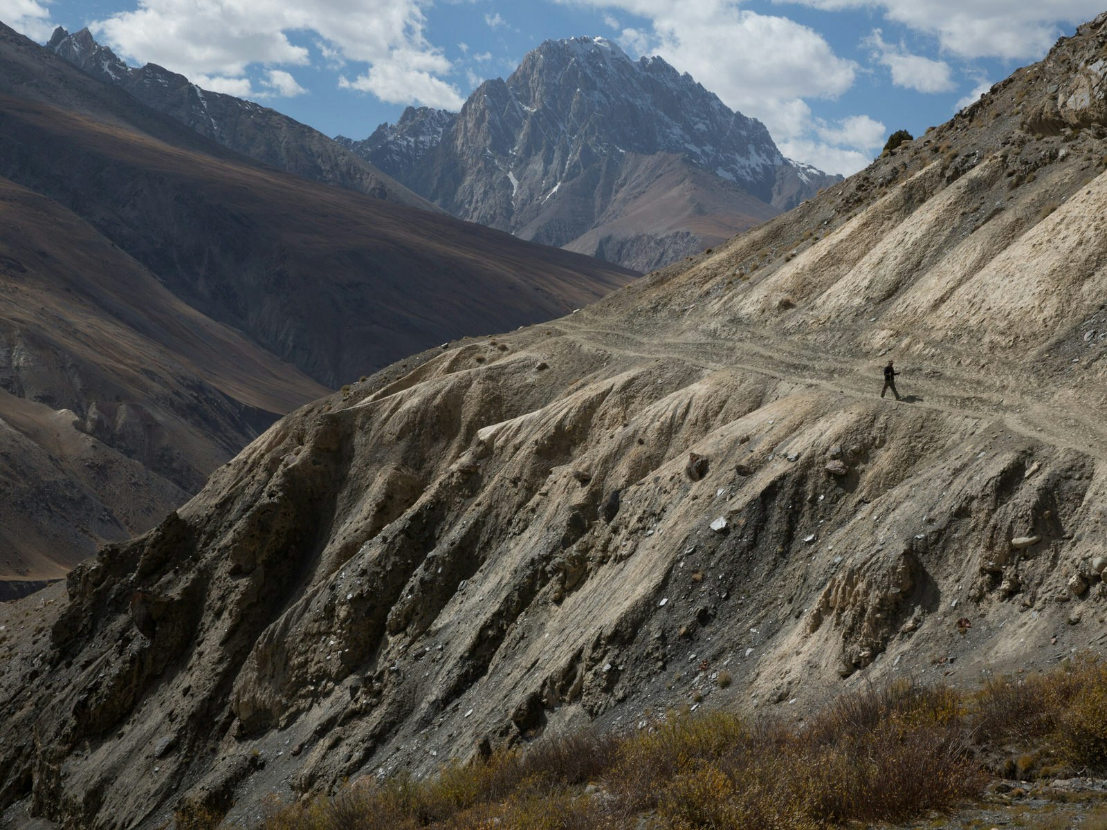 A lone walker treks along a steep mountainside path with tall peaks in the background