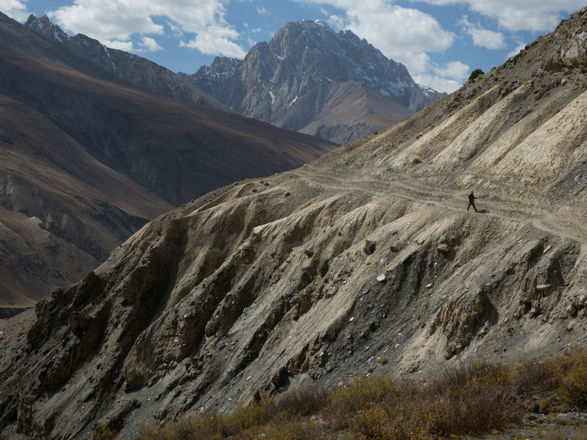 A lone walker treks along a steep mountainside path with tall peaks in the background