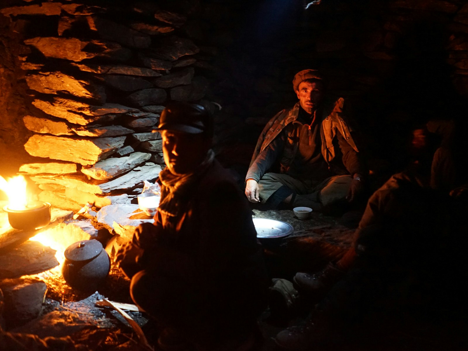 Night in the stone shelter, with a fire to the left and men in traditional hats seated half in the dark