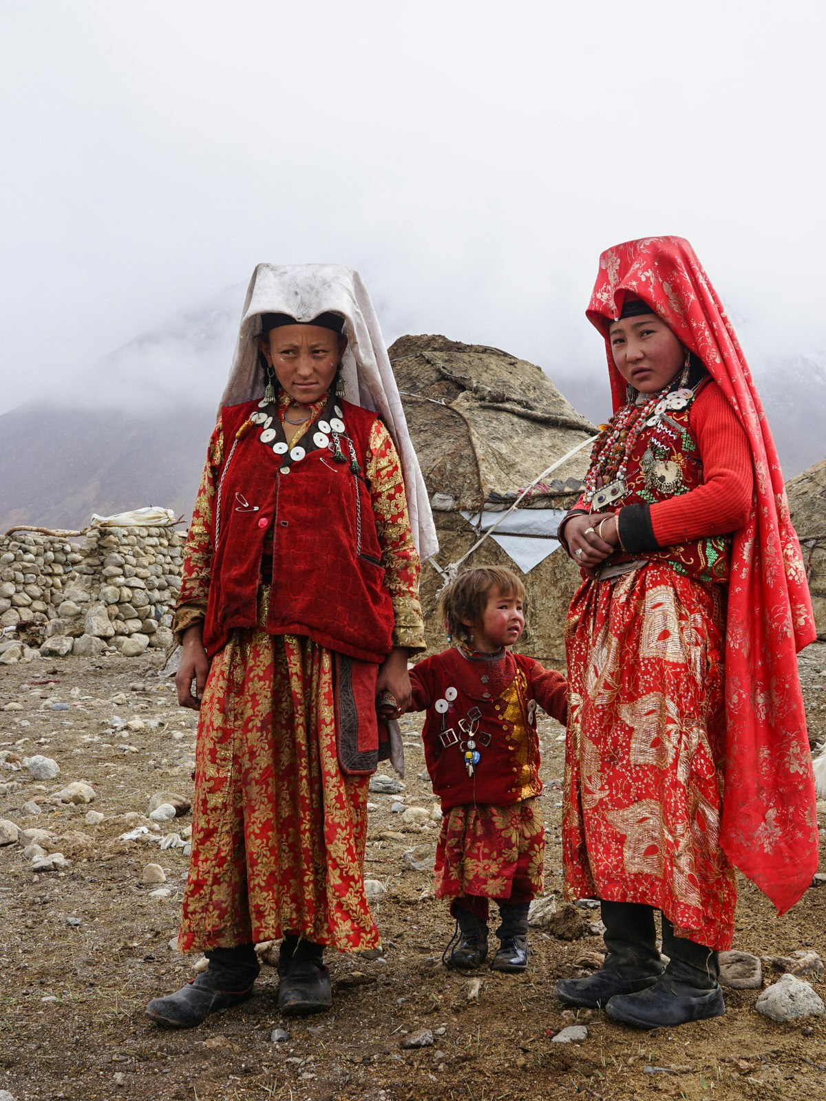 Two young woman dressed in long red embroidered robes and headgear, stand with a baby in front of a yurt tent