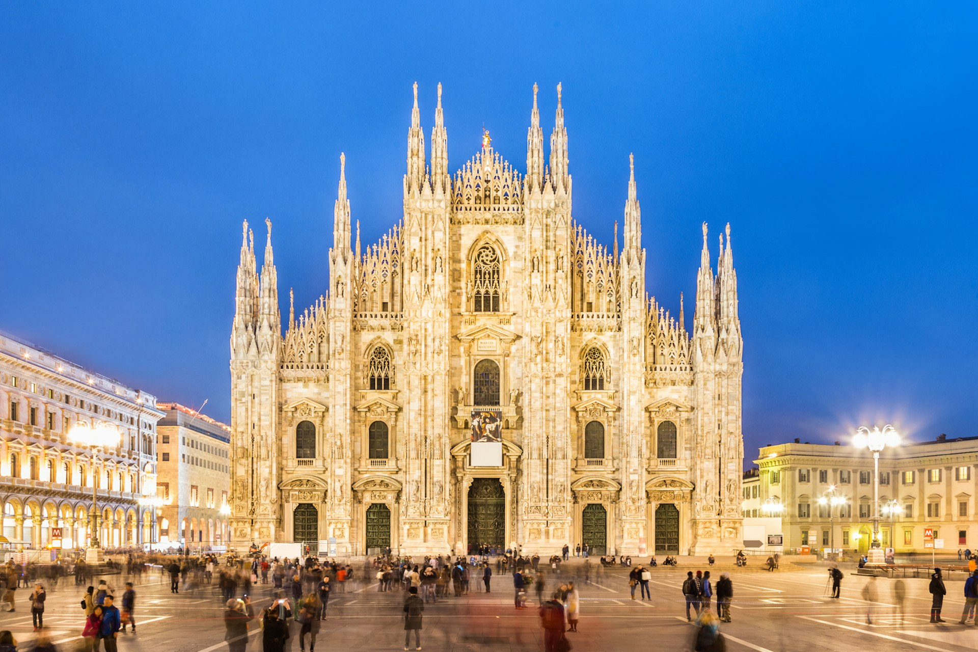 One of the best free things to do in MIlan is admire the Gothic Milan Cathedral; here it is pictured as night falls; its illuminated white marble facade is adorned with many spires and statues; people fill the piazza in front of the cathedral.