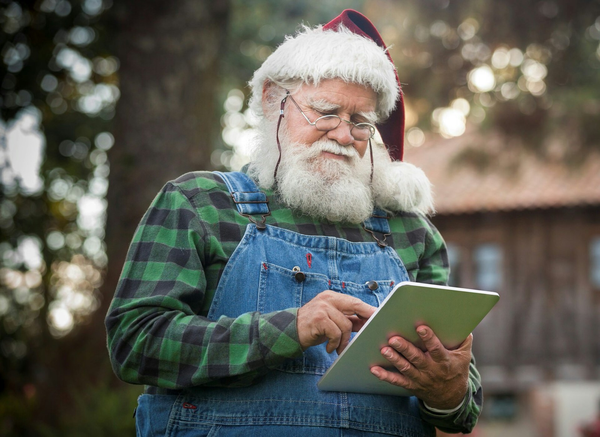 Santa Claus stands in a green check shirt and dungarees using a tablet computer 