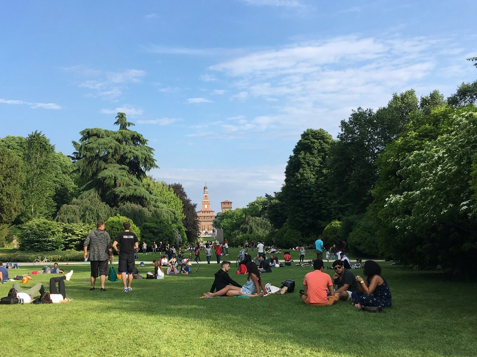 Locals hanging out on the grass in Parco Sempione; there are trees and bushes beyond them, with the imposing red-brick Castello Sforzesco visible further on. 