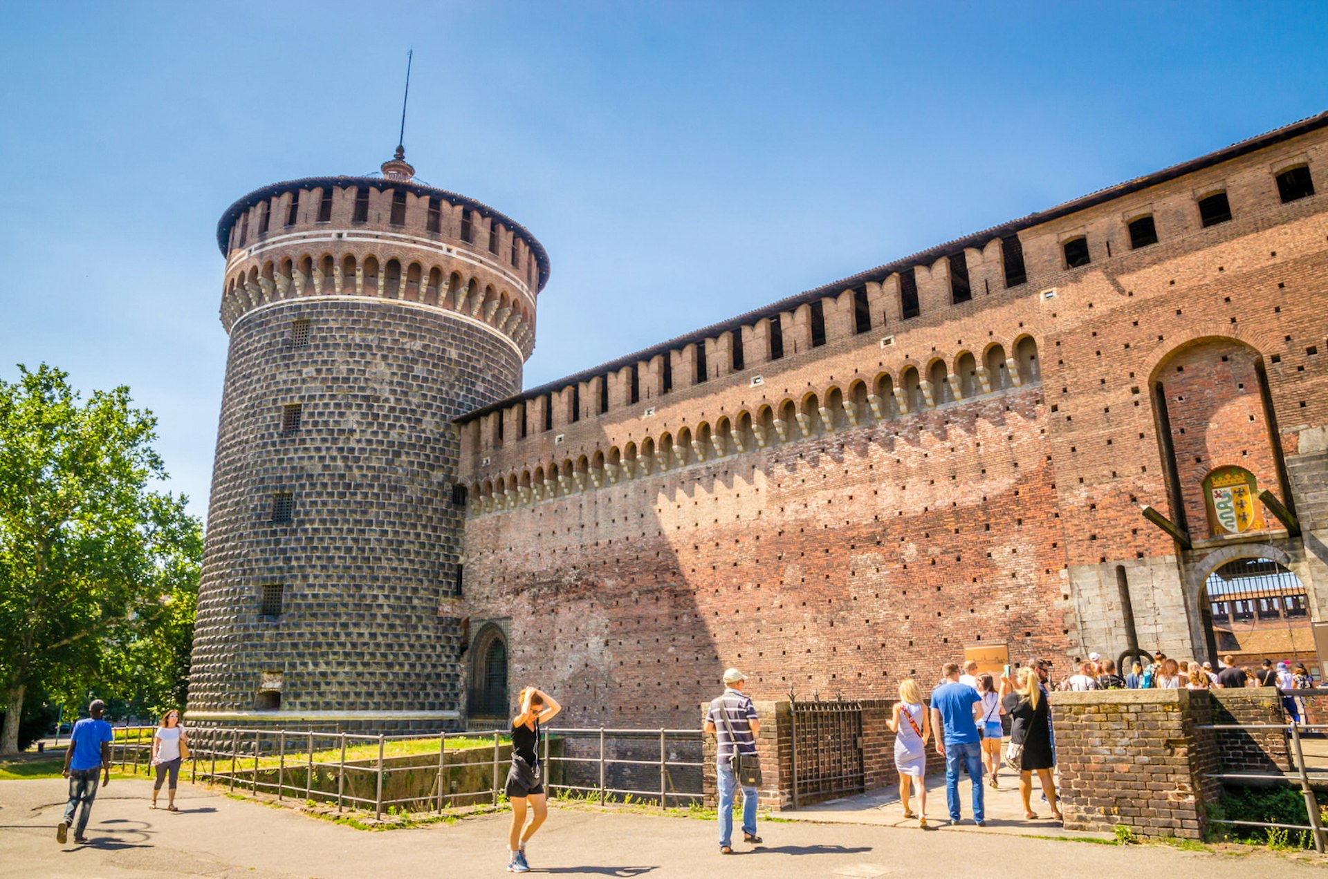 The Sforza Castle, a red-brick fortress with a tower; visitors are walking over a bridge across a moat into the castle.