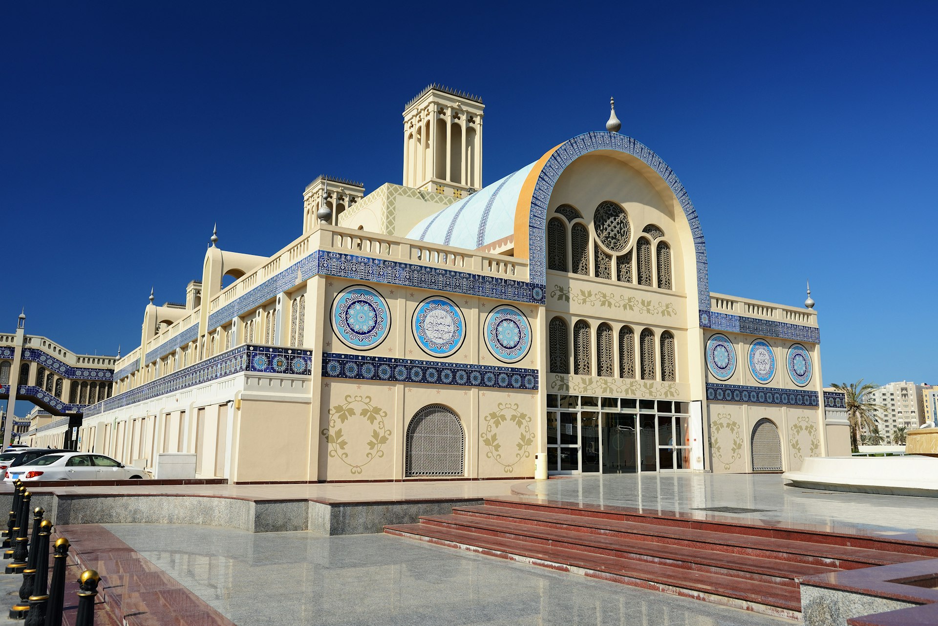Under a cobalt-blue sky sits a large ornate building with a long cylindrical roof with several square towers; the outside is dominated by a creamy yellow colour, with blue accents.