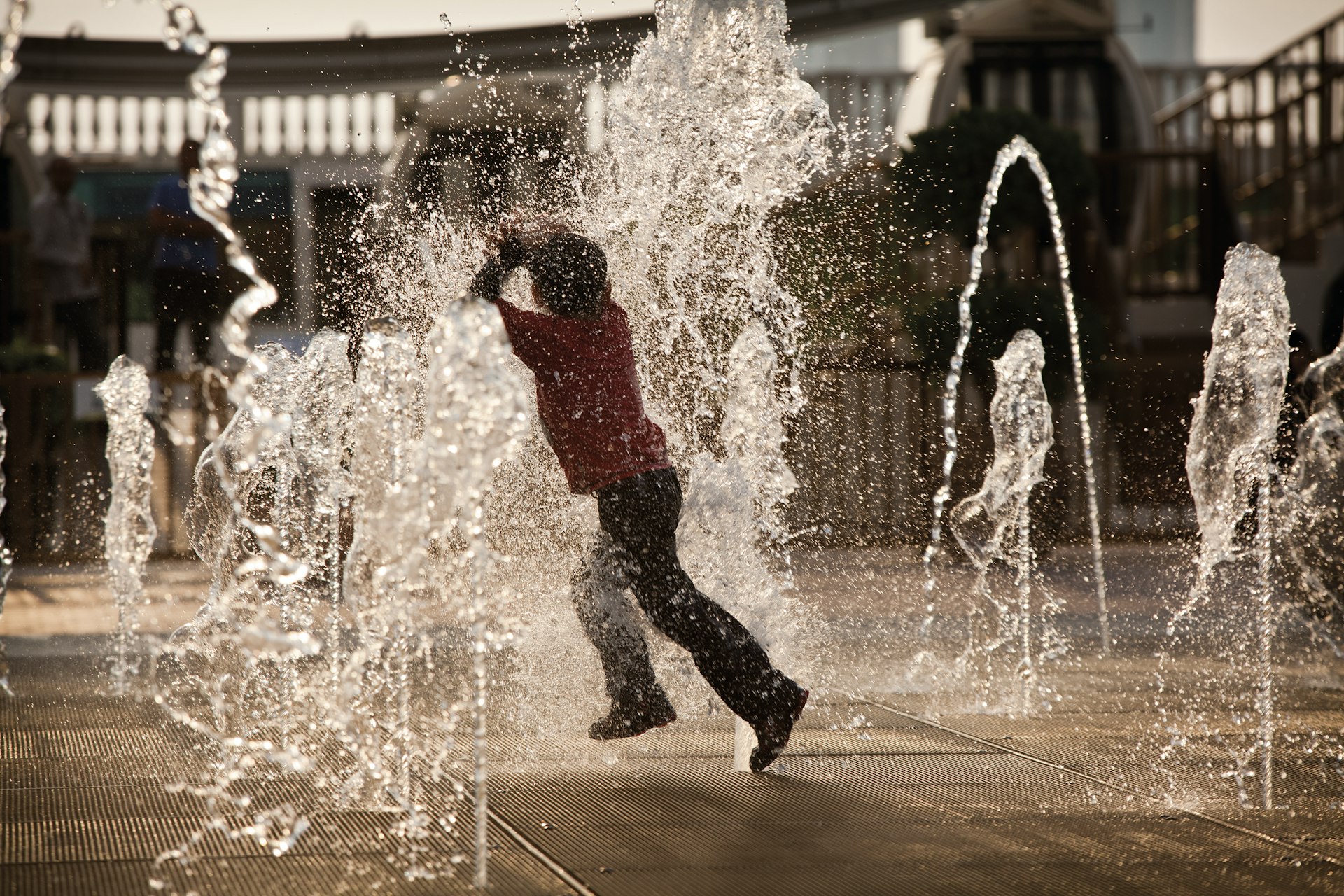 A child leaps through a series of water jets that are firing water up into the air from the pavement.