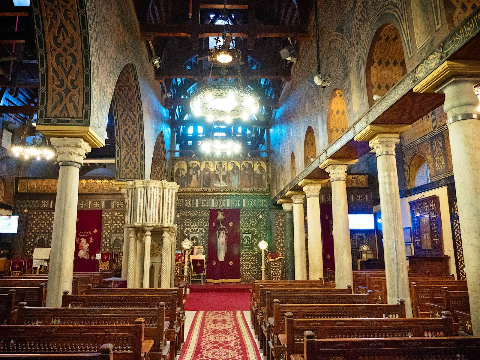 The church interior is dominated by gold patterns on the walls, contrasting with a predominately red carpet and dark wood chairs. Two large circular lights hang down above the central aisle.