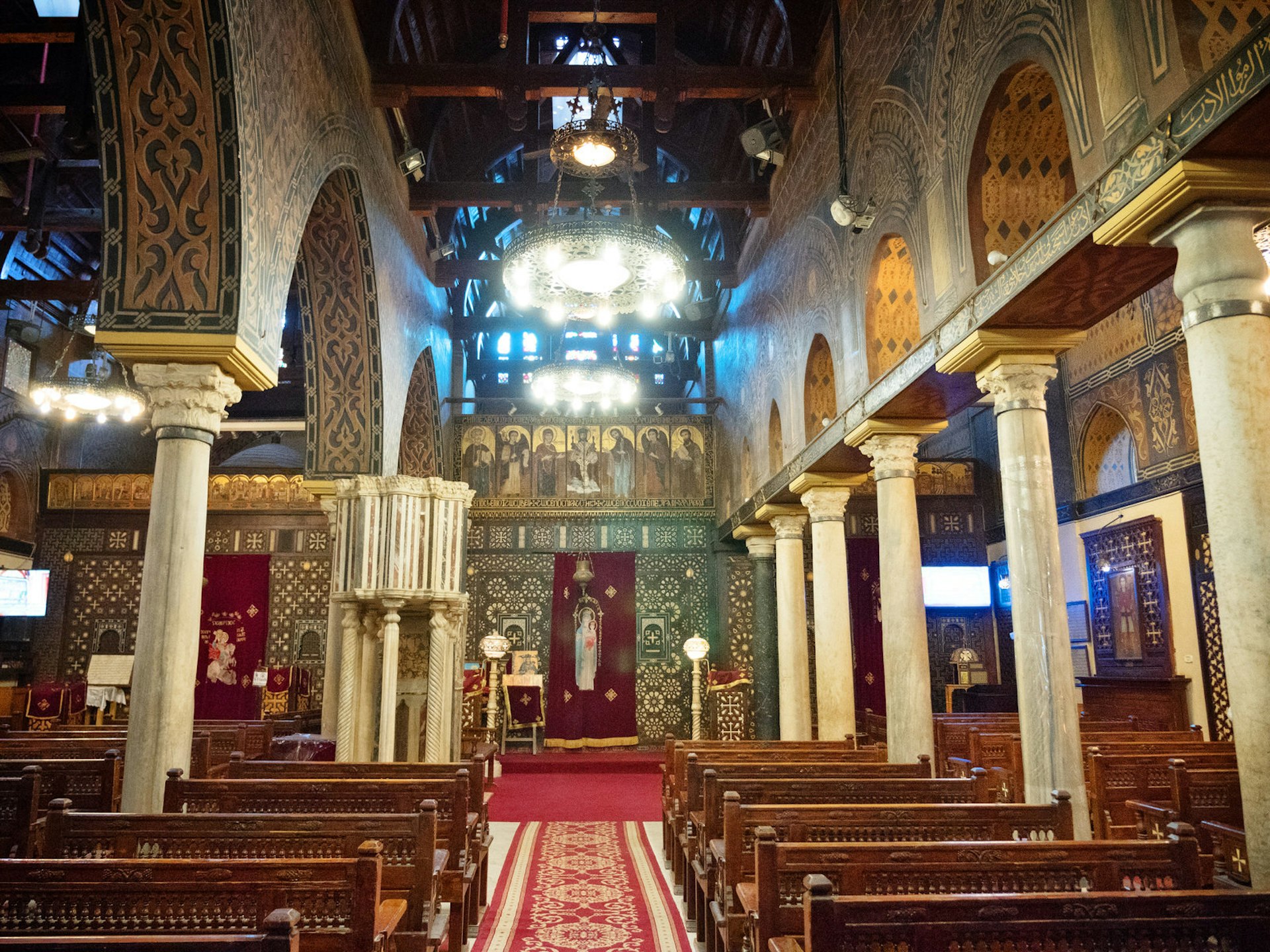 The church interior is dominated by gold patterns on the walls, contrasting with a predominately red carpet and dark wood chairs. Two large circular lights hang down above the central aisle.
