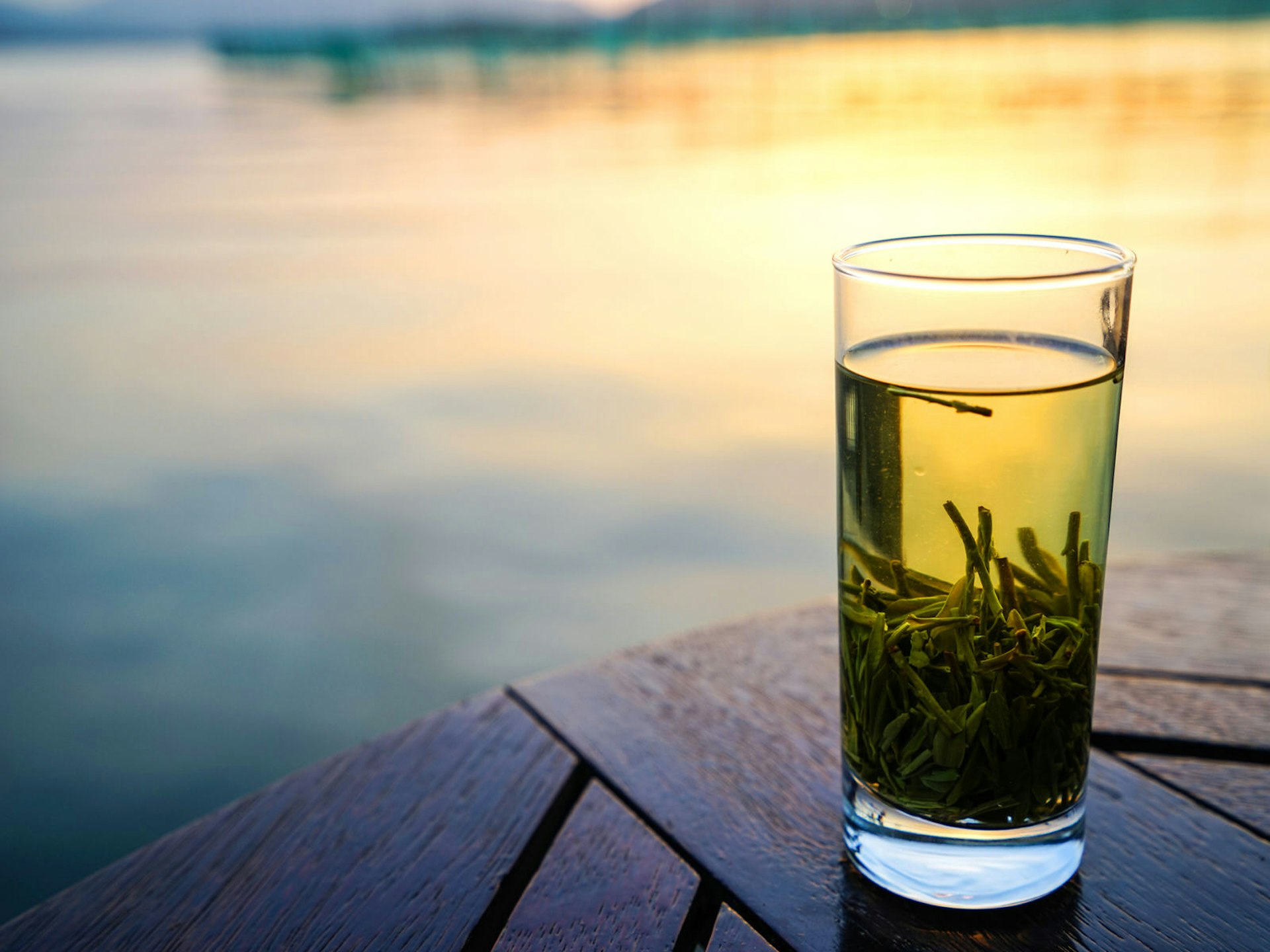 A cup of green tea settled in a tall, clear glass, with sunset reflecting in a pond in the background.