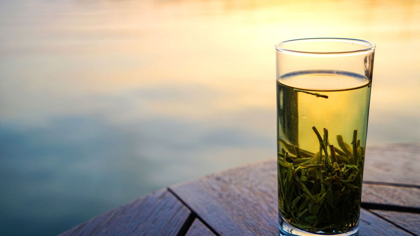 A cup of green tea settled in a tall, clear glass, with sunset reflecting in a pond in the background.