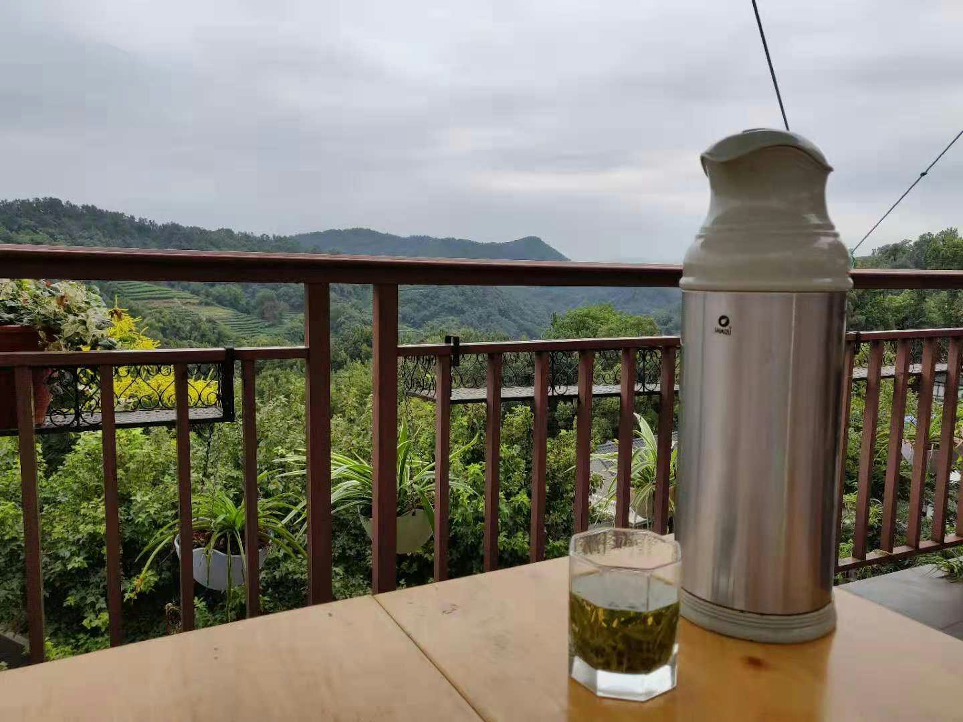 A small glass of tea with large silver flask of hot water on a balcony table overlooking Hangzhou's tea fields
