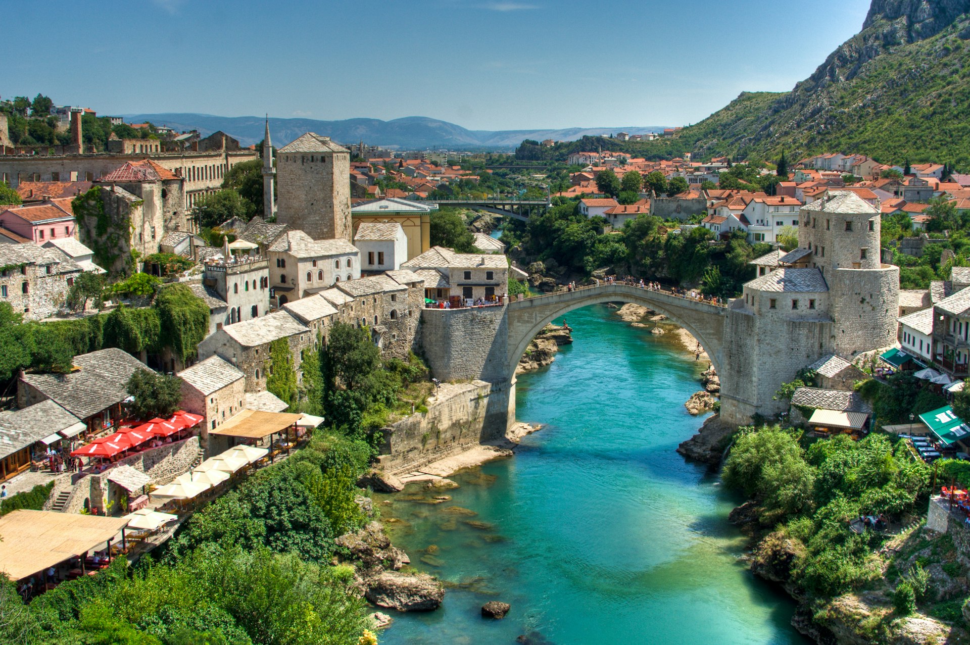 A turquoise river flows underneath an arched bridge, which connects two sides of the city of Mostar