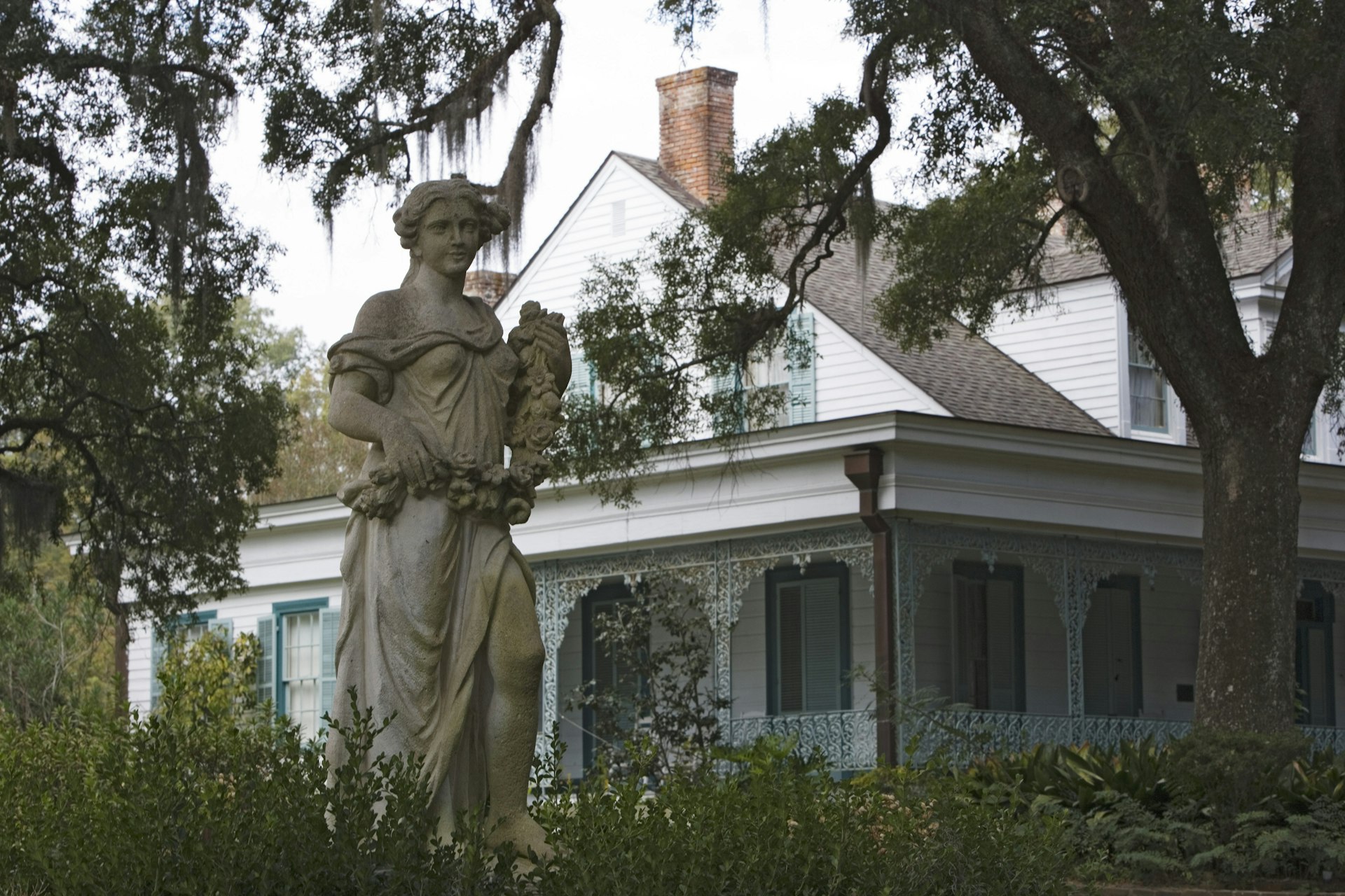 A statue of a woman holding flowers is positioned in front of a white house with light blue shutters on Myrtles Plantation in Louisiana 