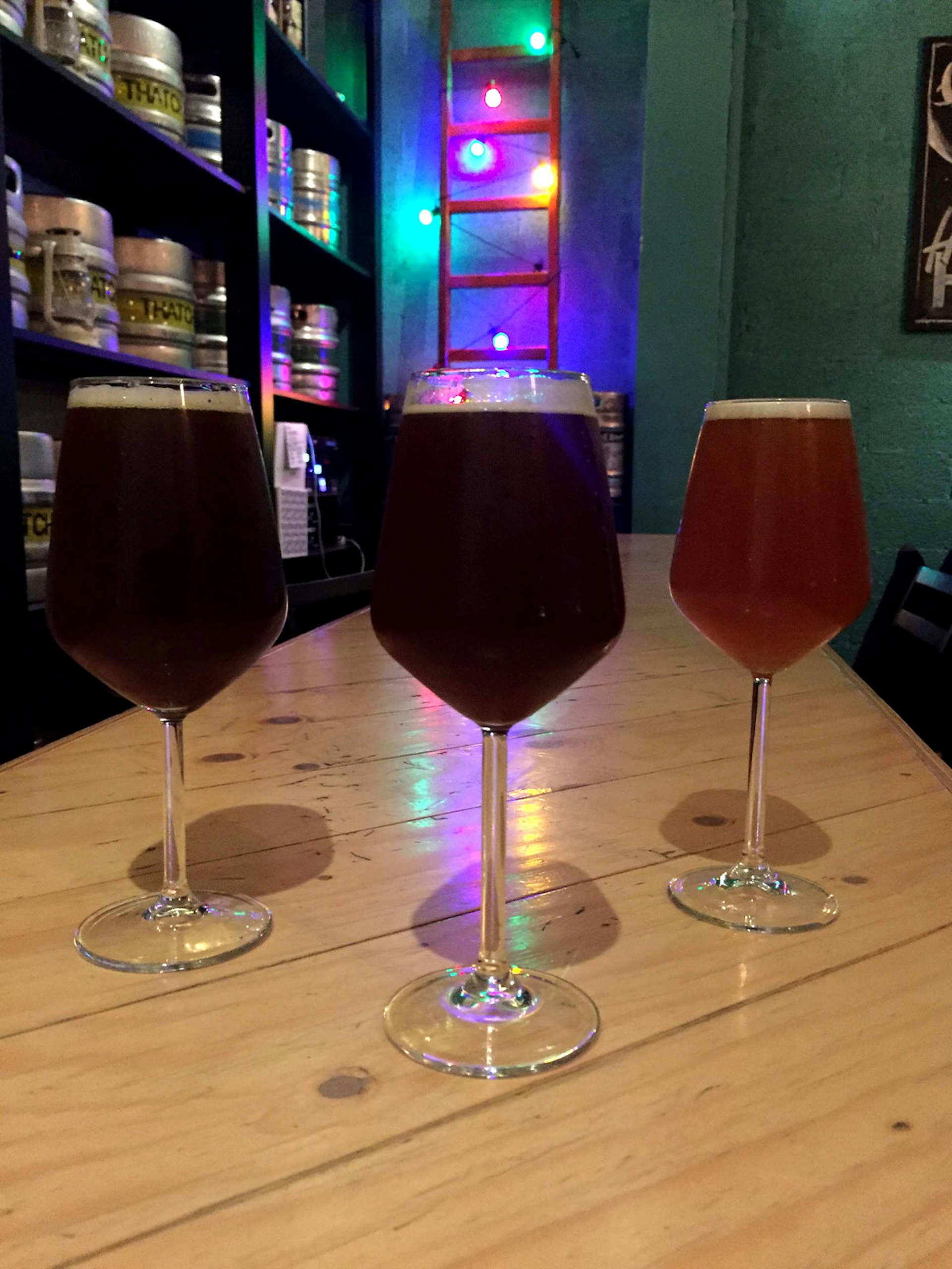 Three glasses of different coloured beers on a table in front of shelves of beer kegs and rainbow lighting