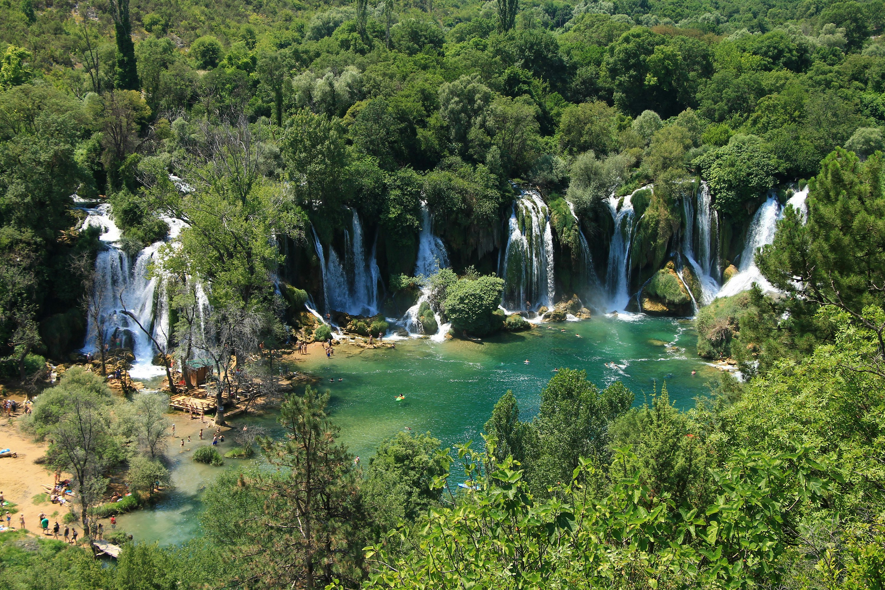 A series of waterfalls flow out of dense forest into a green-blue pool. A few people can be seen on the beach to the left.