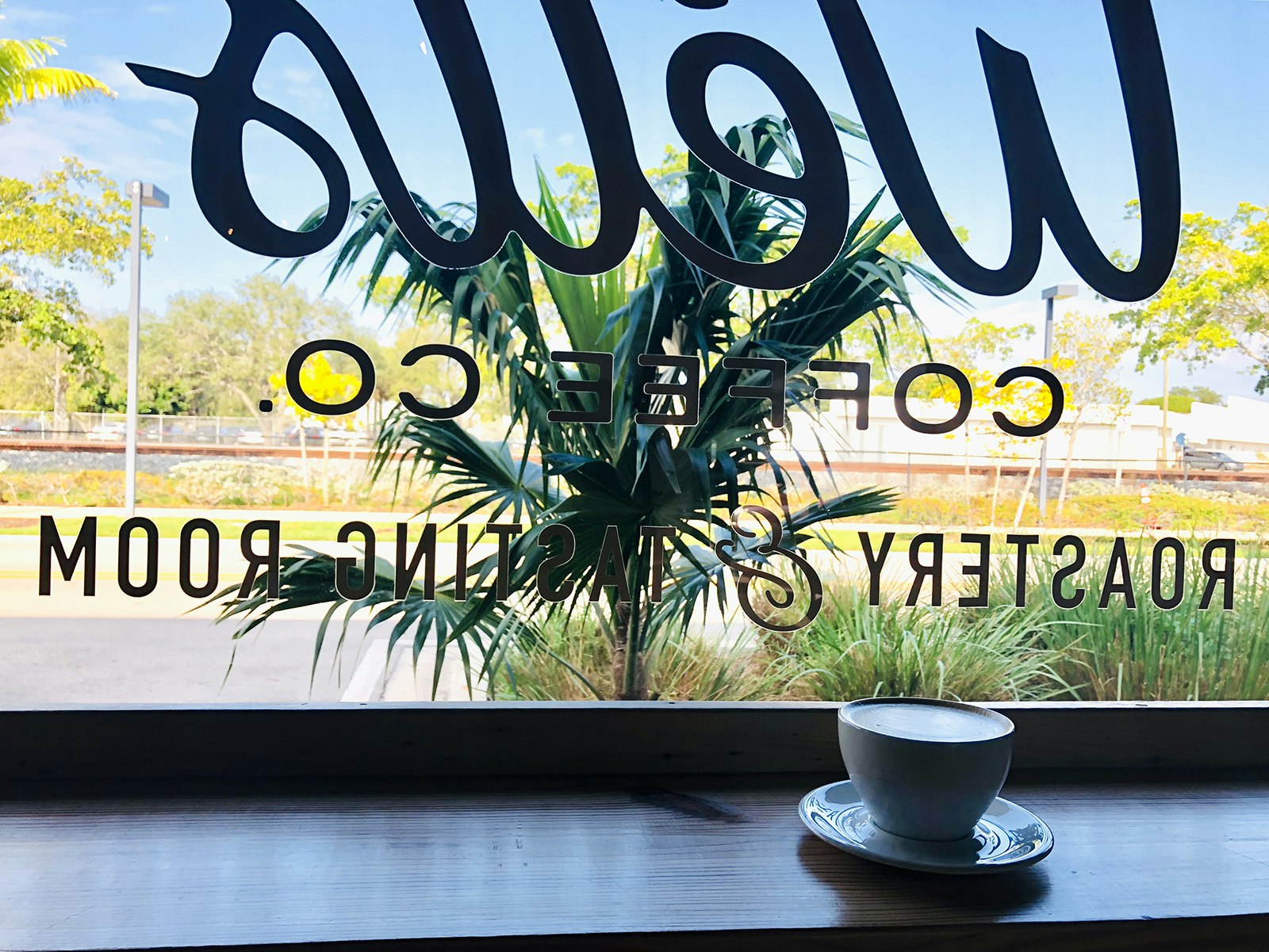 A latte sits on a wooden ledge behind a coffee shop's front window looking out over palm trees and a train track