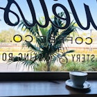 A latte sits on a wooden ledge behind a coffee shop's front window looking out over palm trees and a train track