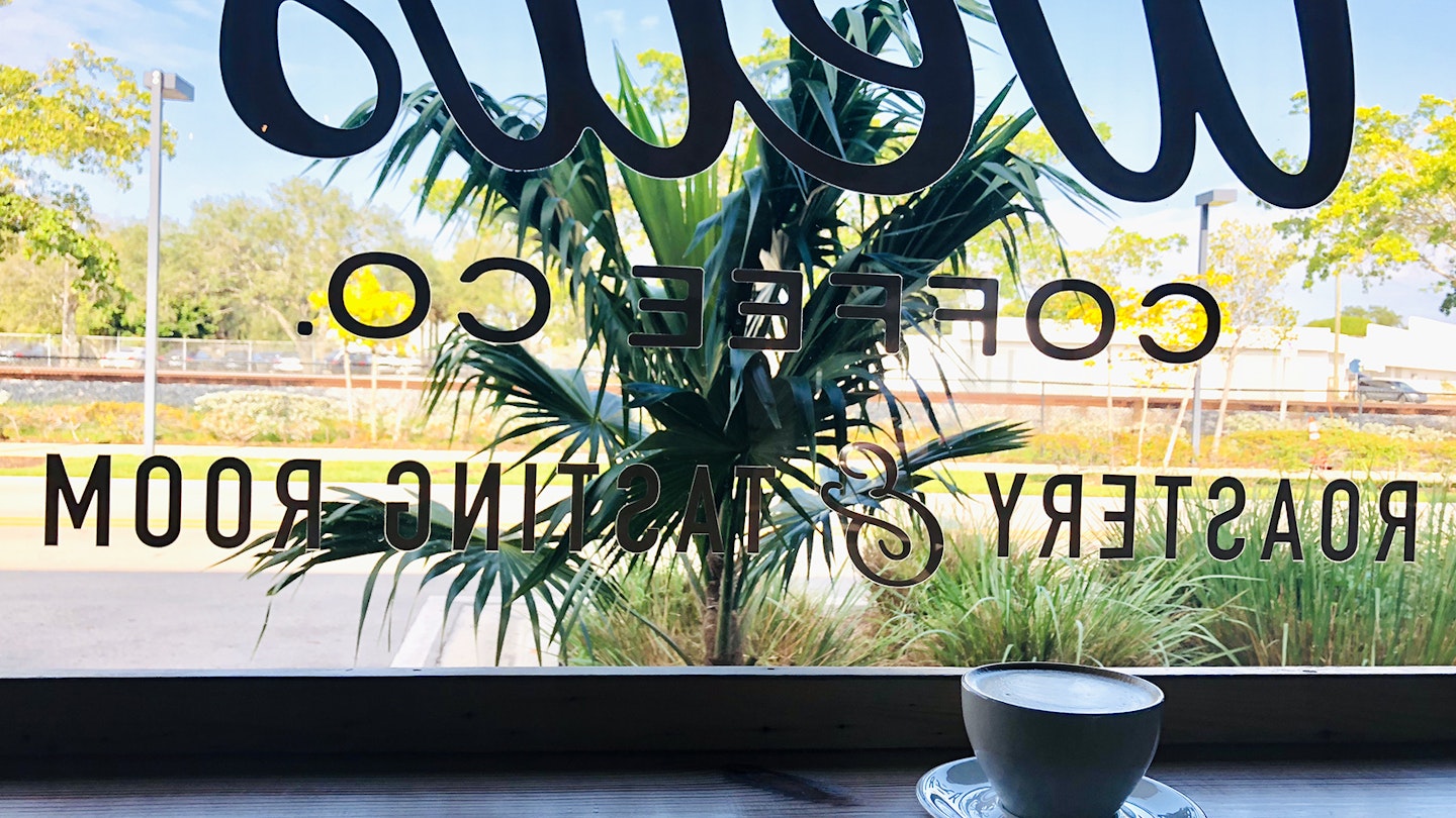 A latte sits on a wooden ledge behind a coffee shop's front window looking out over palm trees and a train track