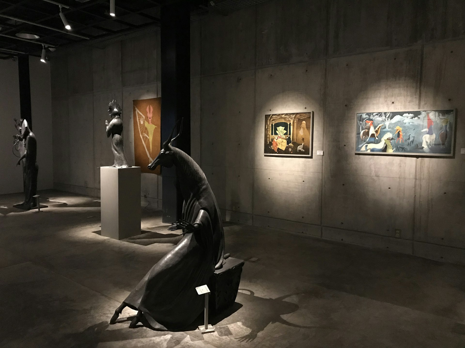 Surrealist paintings hang on the walls of a cement room with bronze sculptures in the foreground