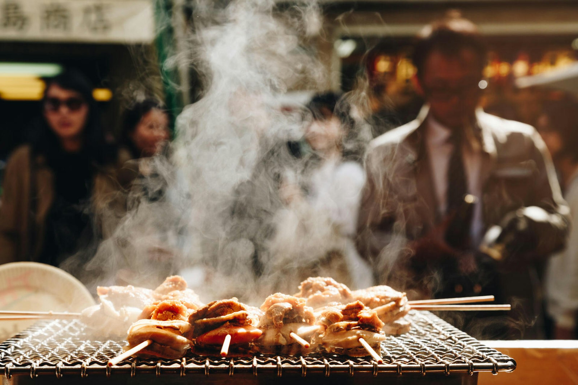 Grilled seafood scallop and sea urchin eggs skewers, with smoke curling above them, lie on a metal grill at a Japanese street food stall at Tokyo's Tsukiji Fish Market