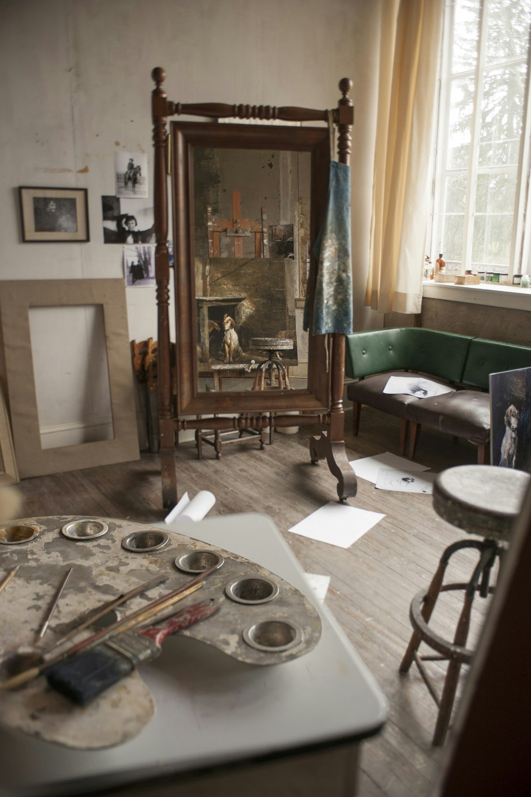 An antique, wood-framed, freestanding mirror in the centre of a room, where pieces of sketch paper are littered across the floor, photographs adorn the walls and a set of oil paints are visible in the foreground, at the studio of painted Andrew Wyeth in Pennsylvania
