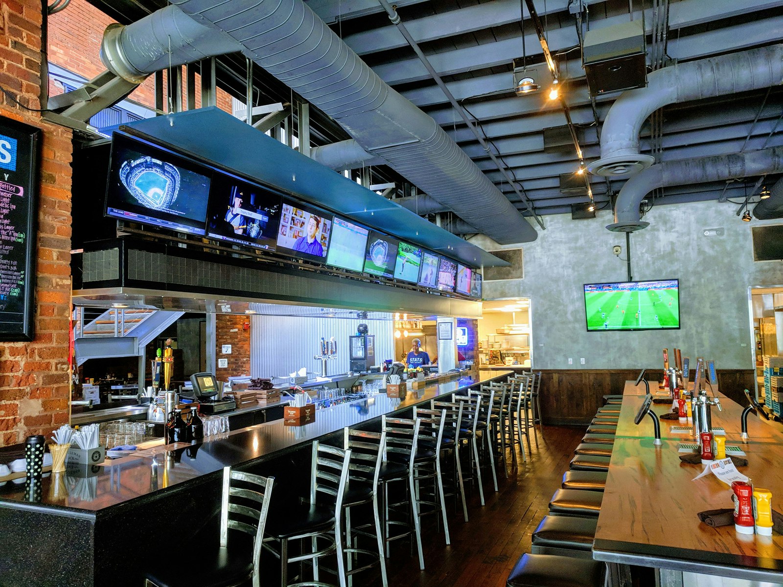 Interior shot of a long bar with a black top and silver bar-height chairs lined up, facing a row of TVs