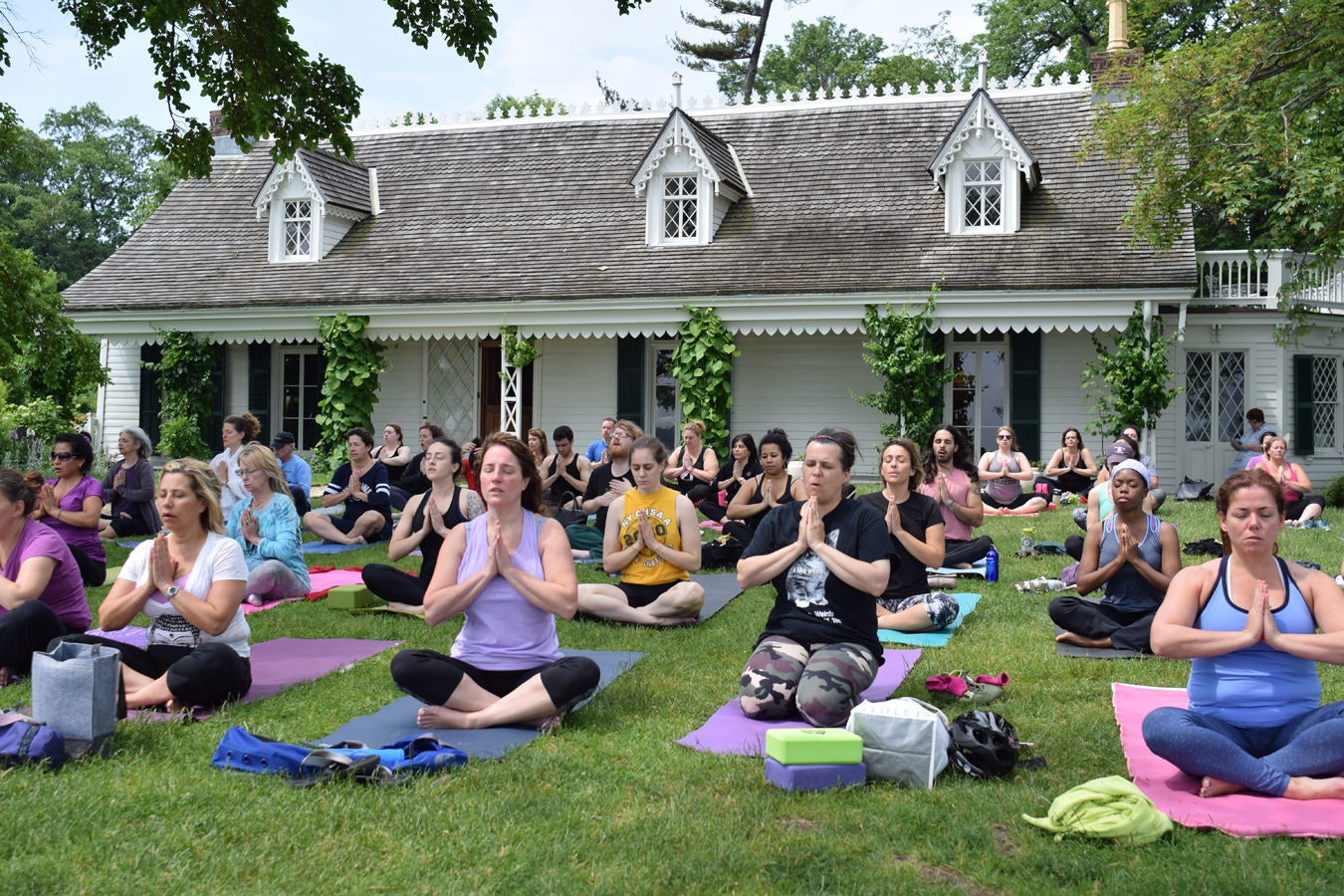 On a grassy lawn in front of the attractive, ivy-wreathed Alice Austen House on Staten Island in New York, yoga practitioners are cross-legged with their hands pressed together