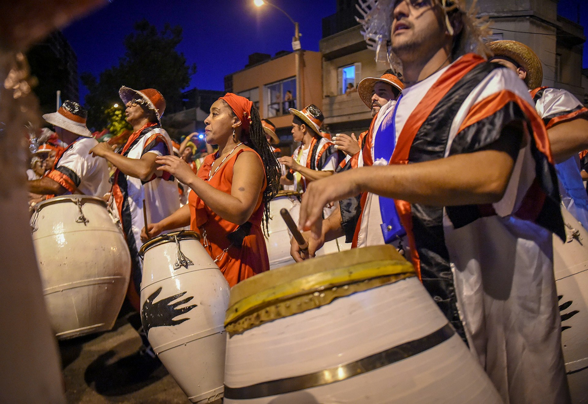 A group of people clad in orange, black and white robes play large white drums