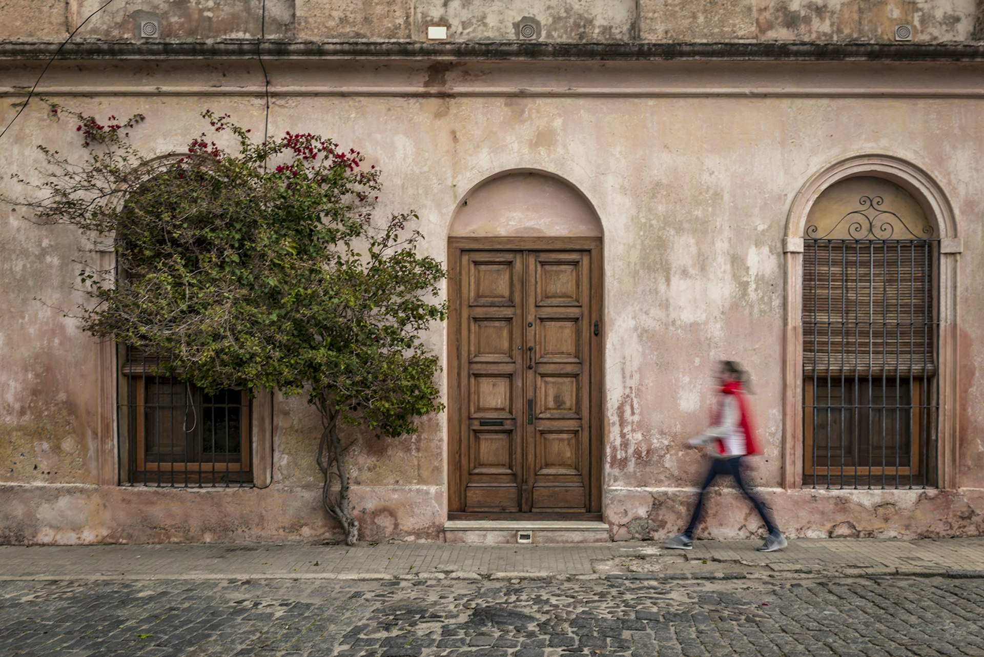 A long exposure of a woman wearing a red scarf walking in front of a worn, pink building with a large wooden door and a ficus tree growing up the wall