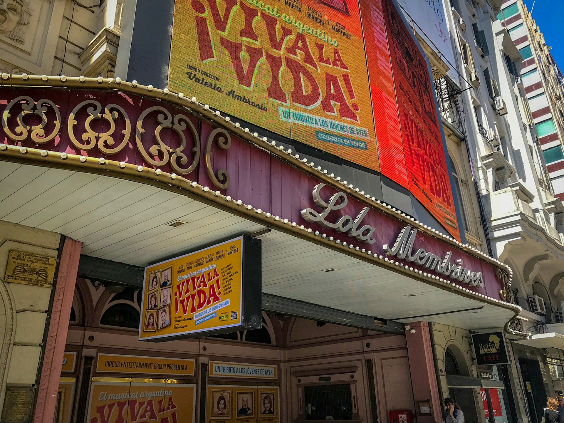 A purple marquee of a theater on Avenida Corrientes with its name written in cursive silver script; above is a billboard advertising a show titled "Viva la Vida" 