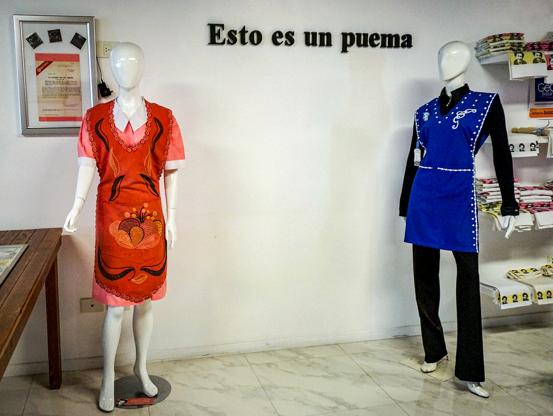 Two mannequins wearing a blue and orange apron, standing against a white wall with "Esto es un puema" written on it 